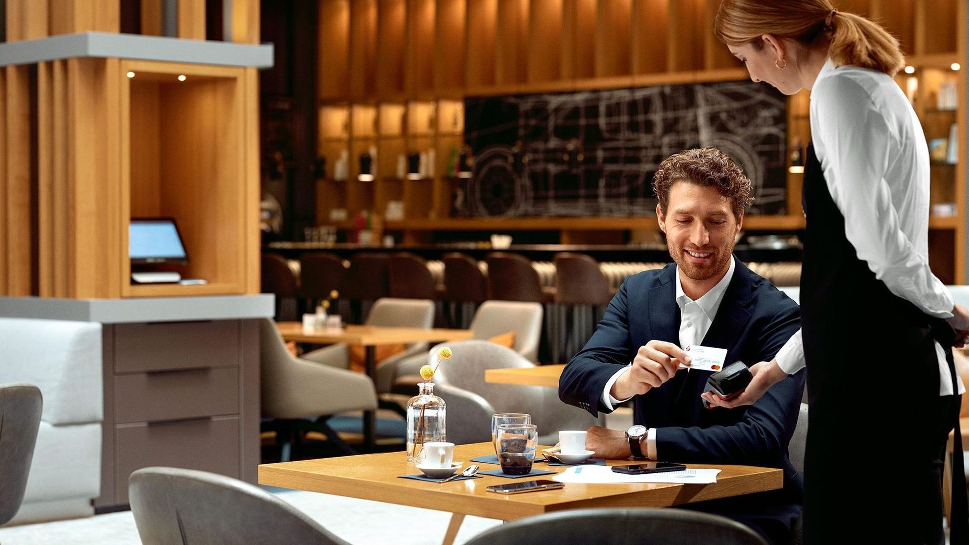 Ein Geschäftsmann mittleren Alters sitzt in einem modernen Restaurant. Er legt lächelnd seine Sparkassenkarte auf ein Kartenlesegerät, welches eine weibliche Servicekraft ihm anreicht.