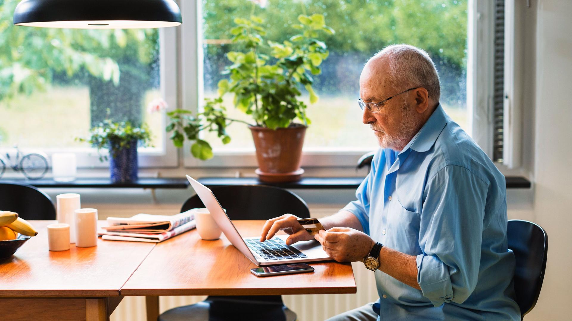 Älterer Mann sitzt mit einem Laptop an einem Tisch und hat eine Kreditkarte in der Hand.