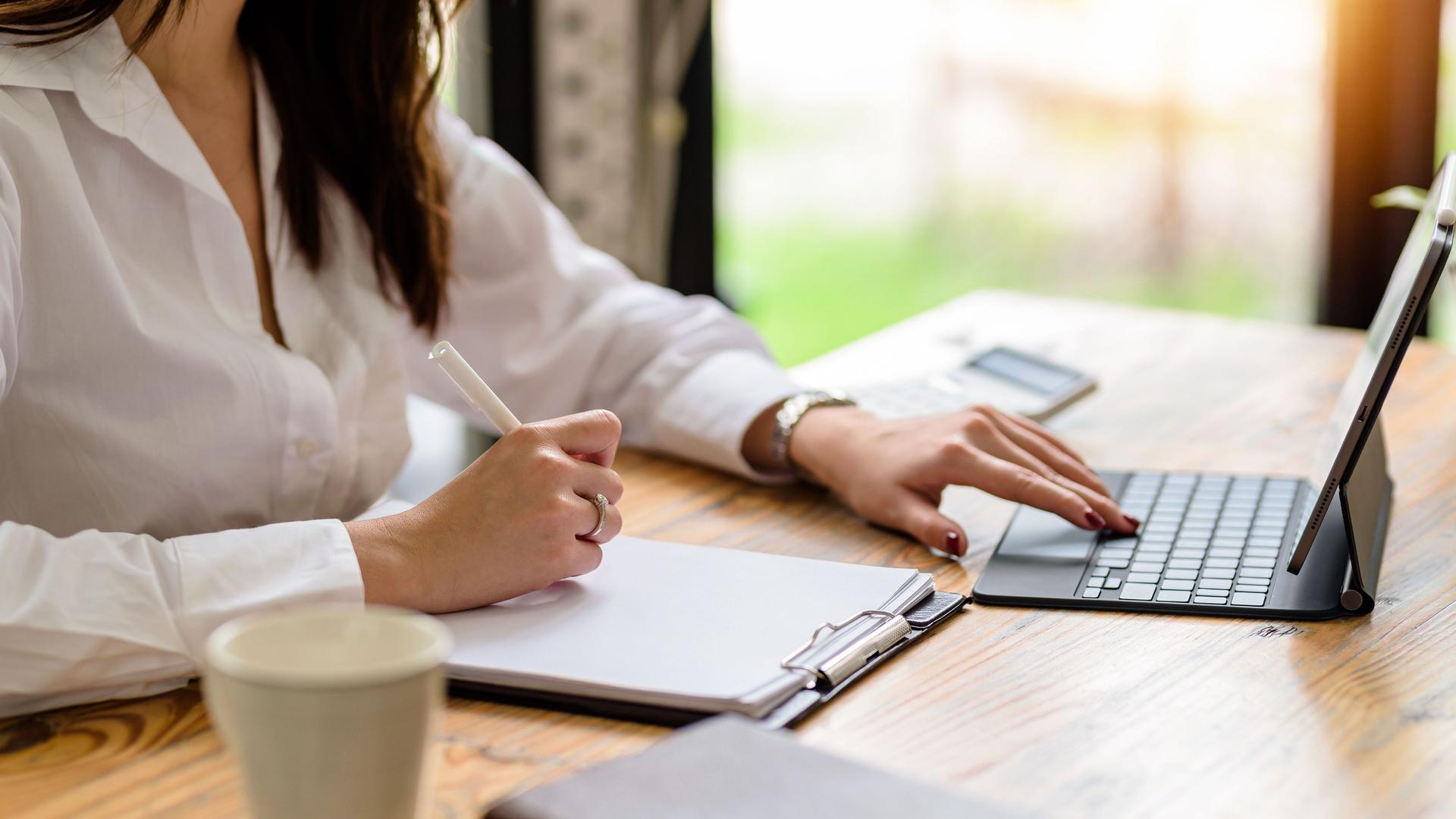 Nahaufnahmebild einer Frau, die auf Notizbuch und Laptop im Büro schreibt bzw. notiert.
