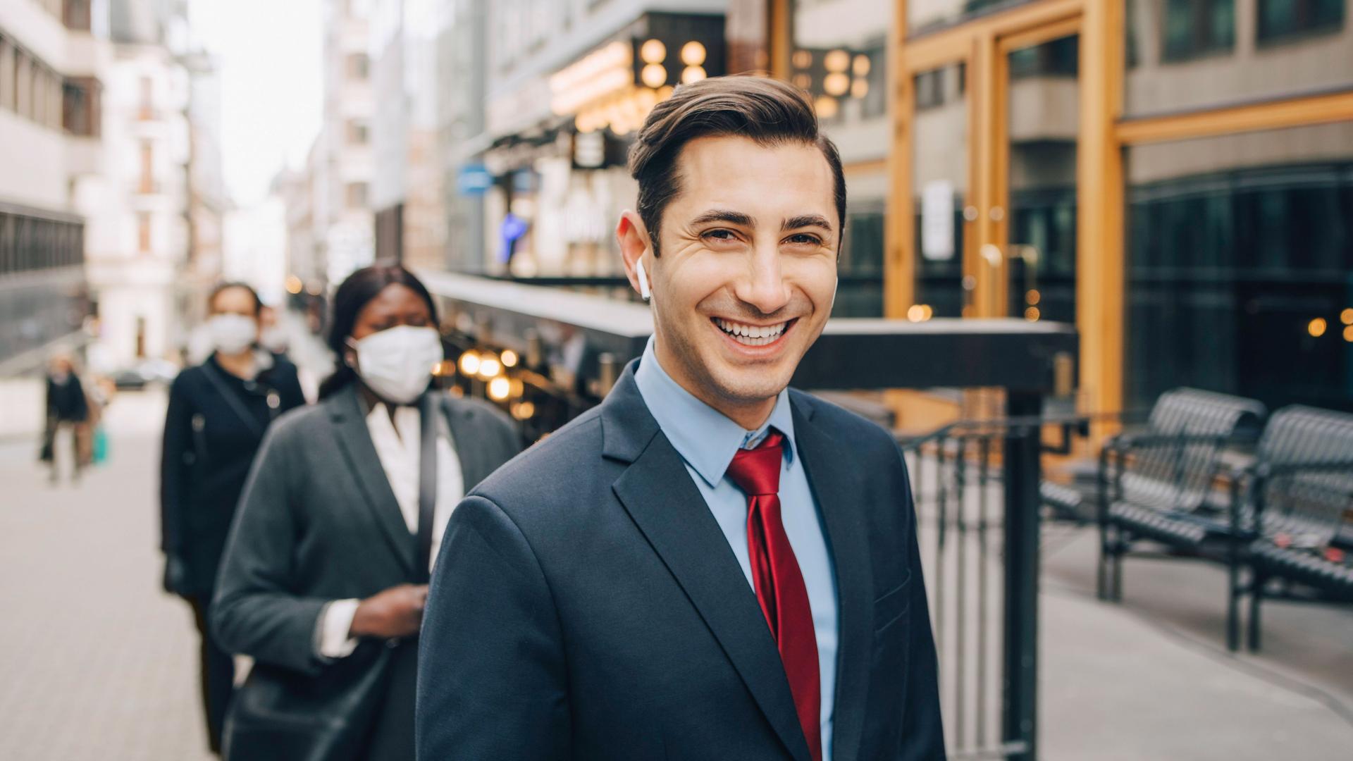 Ein junger Mann in Business-Kleidung läuft auf einer urbanen Straße mit kabellosem Kopfhörer im Ohr und blickt dabei lächelnd in die Kamera, 