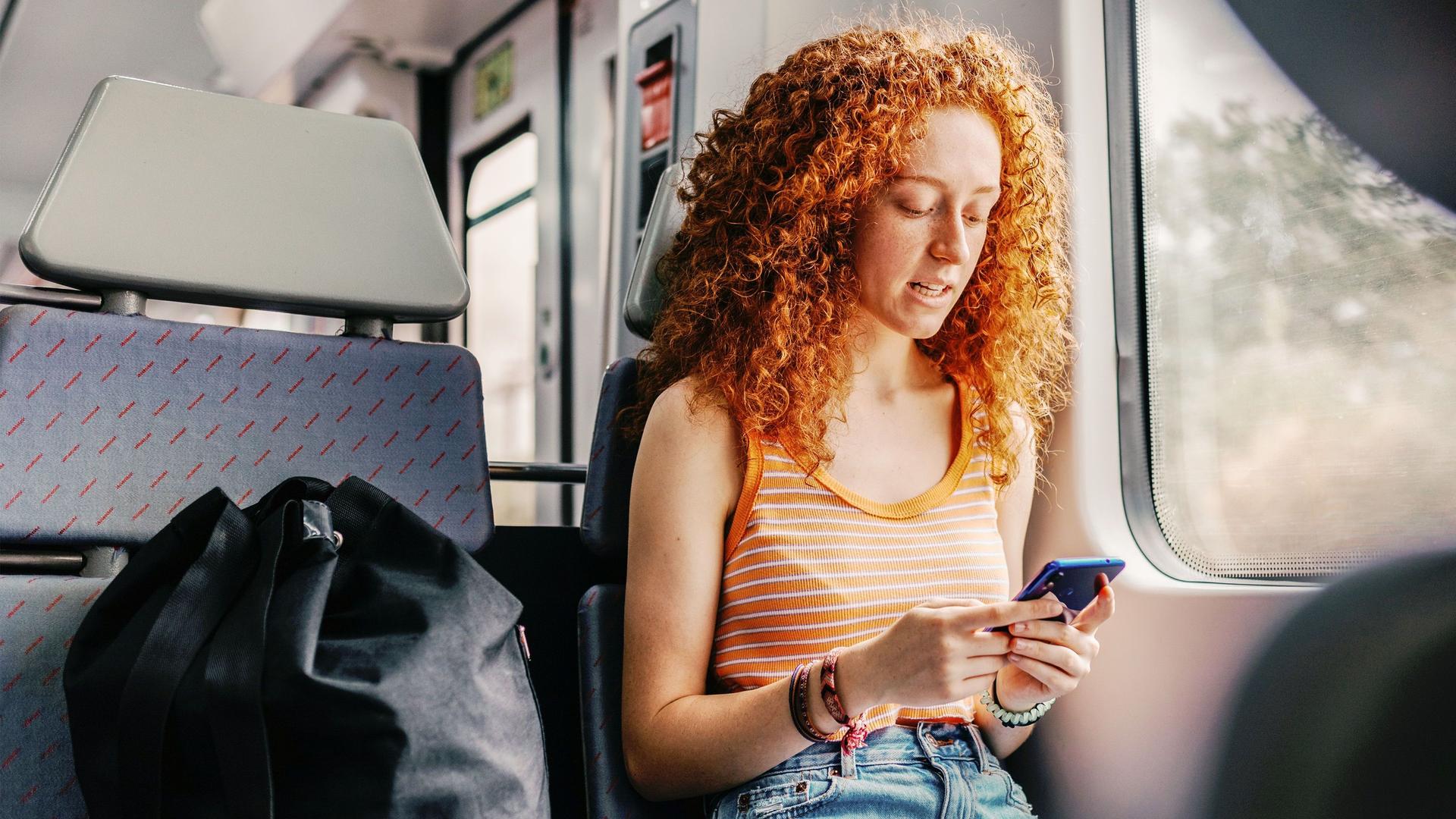 Junge rothaarige Frau sitzt in einem Zug mit Blick auf das Handy in ihrer Hand.