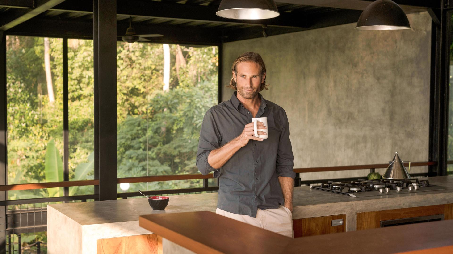 Ein Mann trinkt Kaffee in einer modernen Design-Küche mit Glasfassade, umgeben von einem üppigen tropischen Garten.