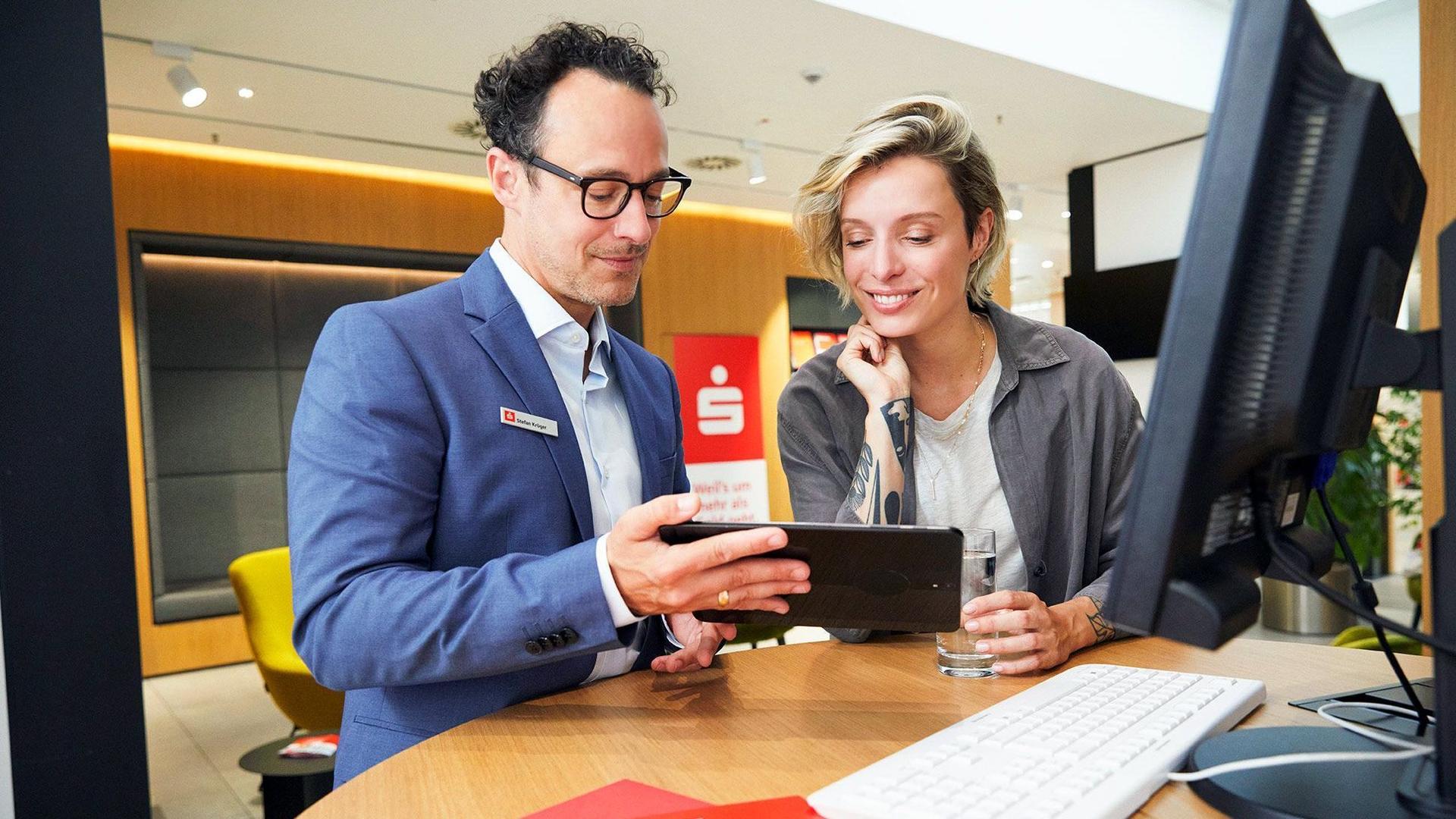Ein Sparkassenberater zeigt einer Kundin in einer modern eingerichteten Filiale etwas auf einem Tabletdisplay. Sie beugt sich interessiert lächelnd nach vorne.