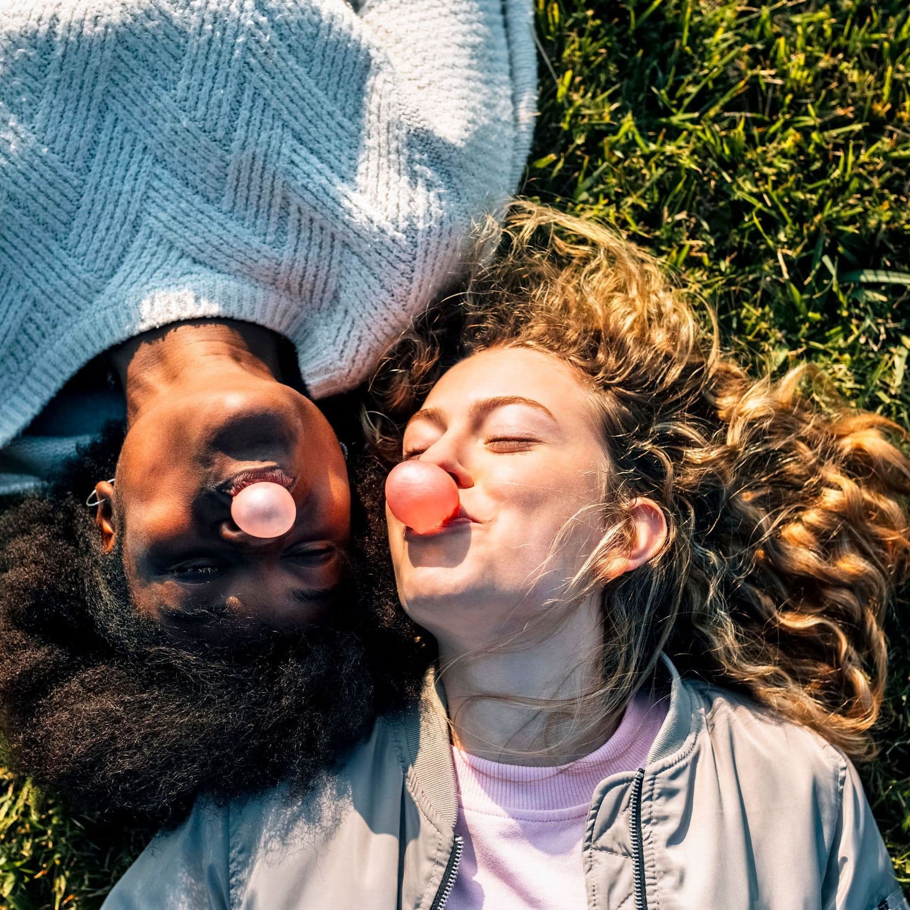 Zwei junge Frauen liegen auf einer grünen  Wiese im Sonnenschein und machen lachend Kaugummiblasen.