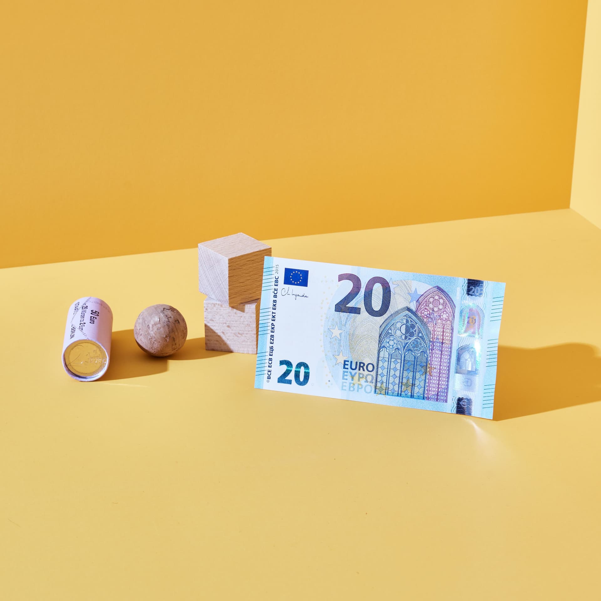 20-Euro-Schein steht aufrecht neben Holzwürfel, Murmel und Münzrolle auf gelbem Untergrund