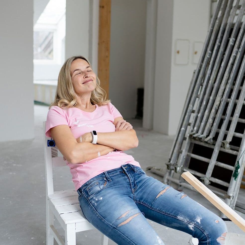 Eine Frau mit blondem Haar sitzt entspannt mit verschränkten Armen und geschlossenen Augen auf einem weißen Stuhl in einem renovierungsbedürftigen Raum. Sie trägt ein rosa T-Shirt, eine Smartwatch und eine zerrissene Jeans. 