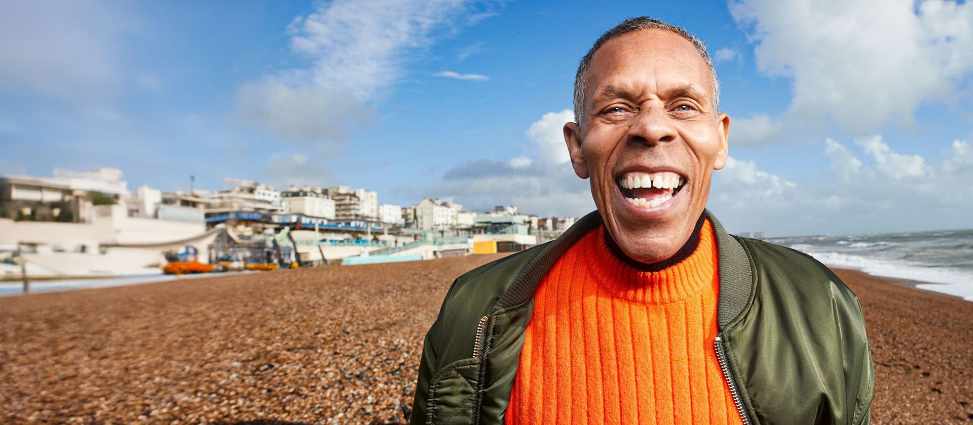 Ein Mann steht lachend an einem Kiesstrand vor einer Stadt am Meer, er trägt einen orangefarbenen Pullover und eine grüne Jacke. Im Hintergrund sind der blaue Himmel und das Meer zu sehen.