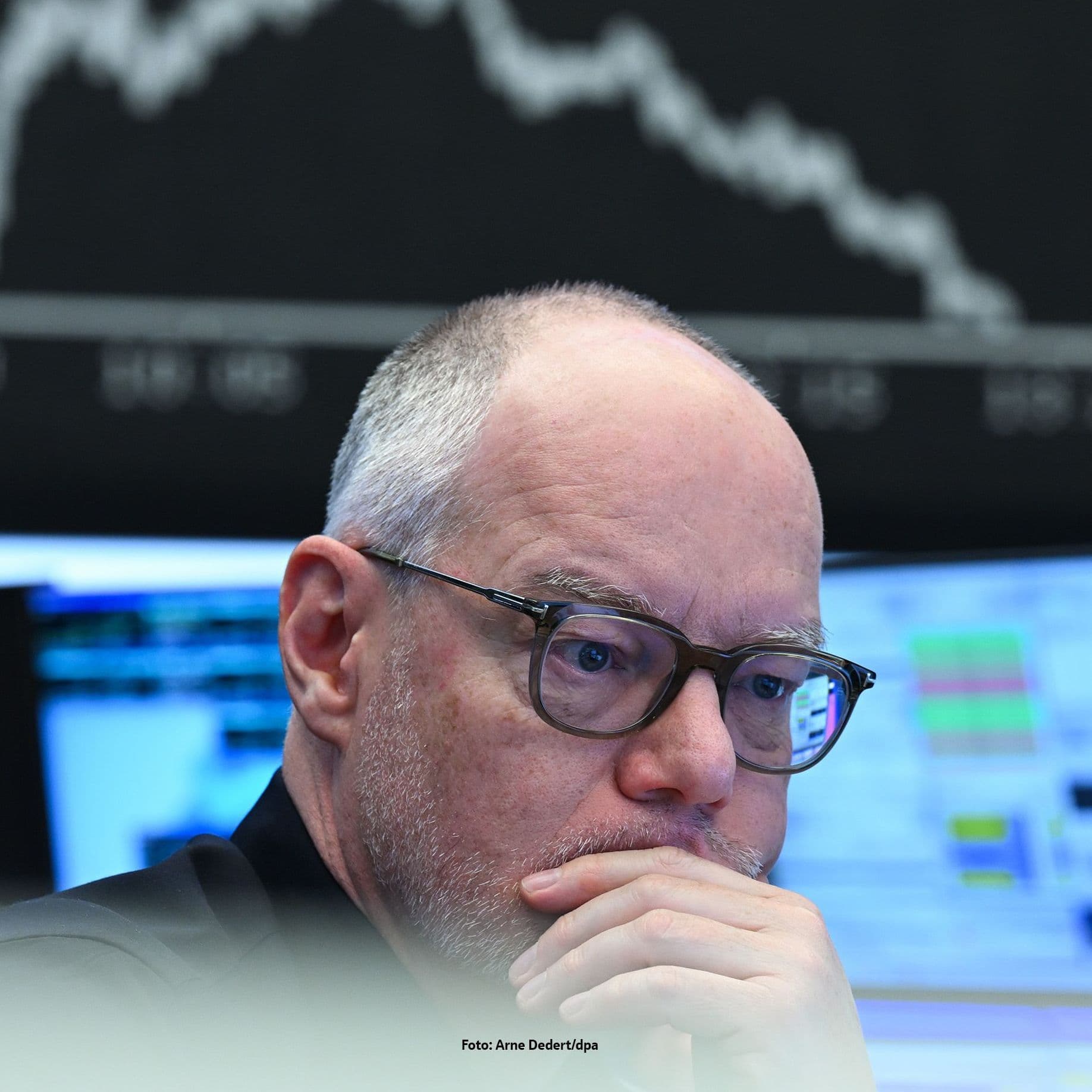 Ein Mann schaut kritisch auf einen Monitor. Im Hintergund ist der fallende Börsengraph zu sehen.