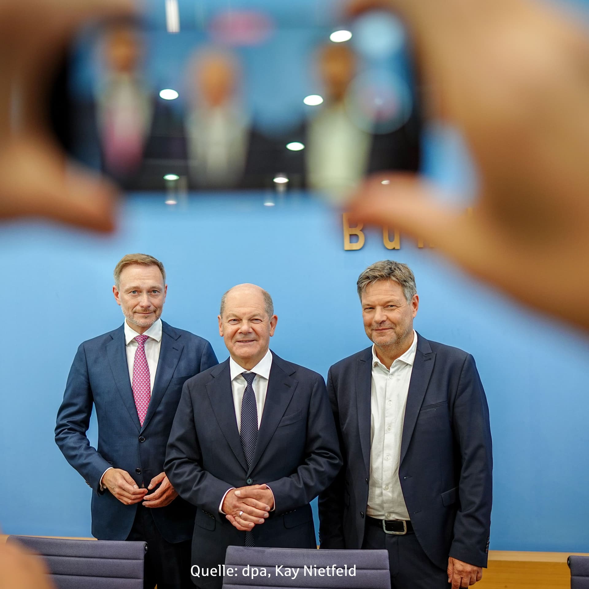 Scholz, Lindner und Habeck stehen vor einer blaugrauen Wand. Im Vordergrund sind Hände mit einem Smartphone zu sehen.