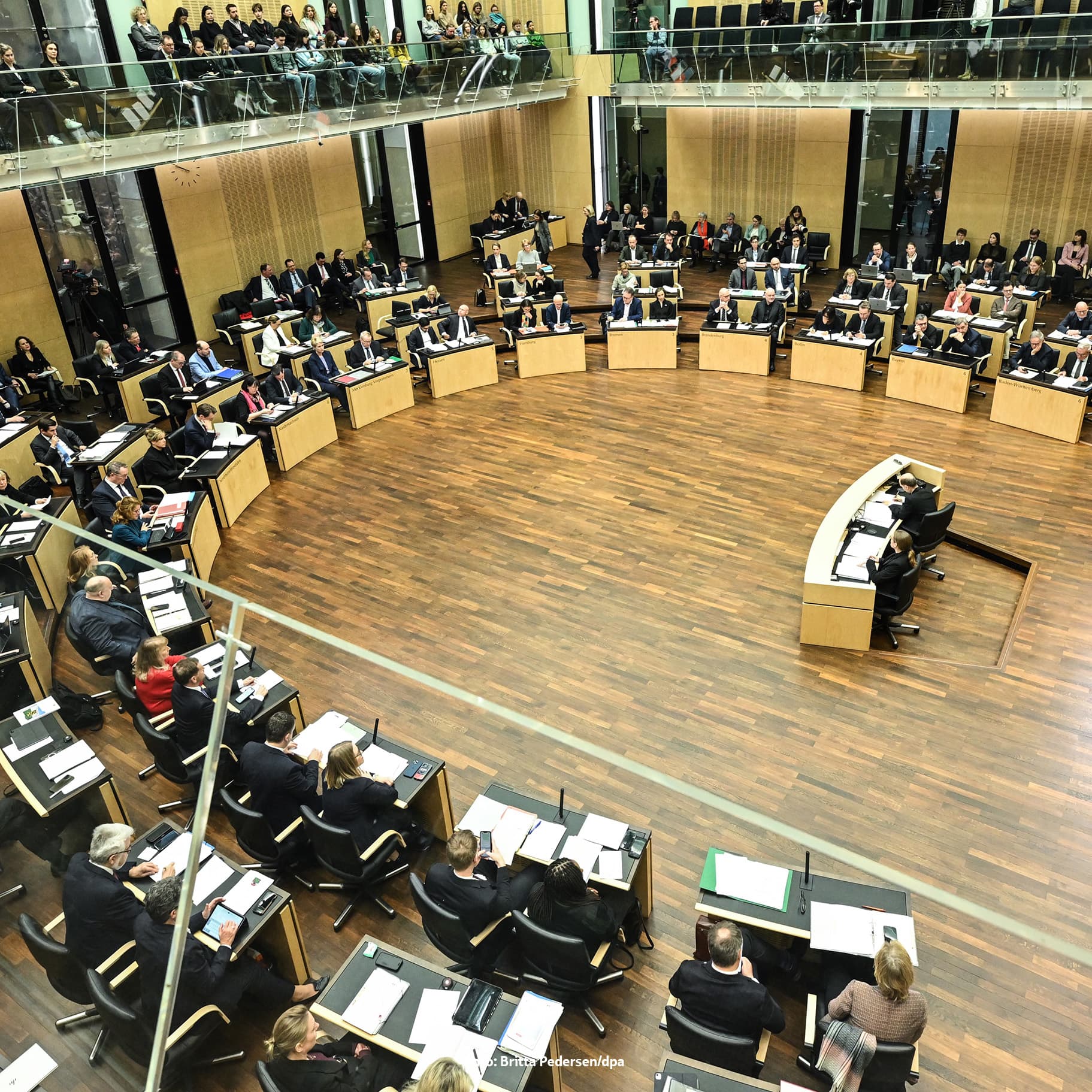 Großer Sitzungssaal in parlamentarischer Runde: Viele Personen sitzen halbkreisförmig an Pulten, ein Redner steht am Rednerpult; Zuschauer auf Galerien im oberen Bereich