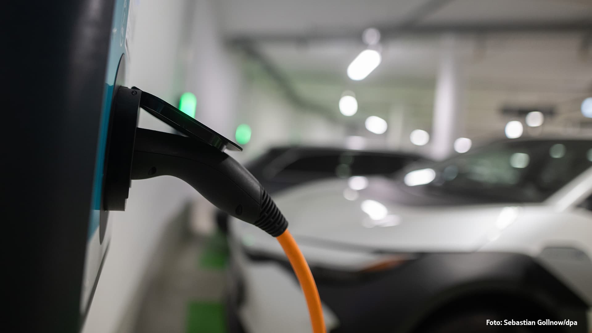 Nahaufnahme eines orangefarbenen Ladekabels, das an eine Ladesäule für Elektroautos angeschlossen ist, mit einem unscharfen Auto und Lichtern im Hintergrund.