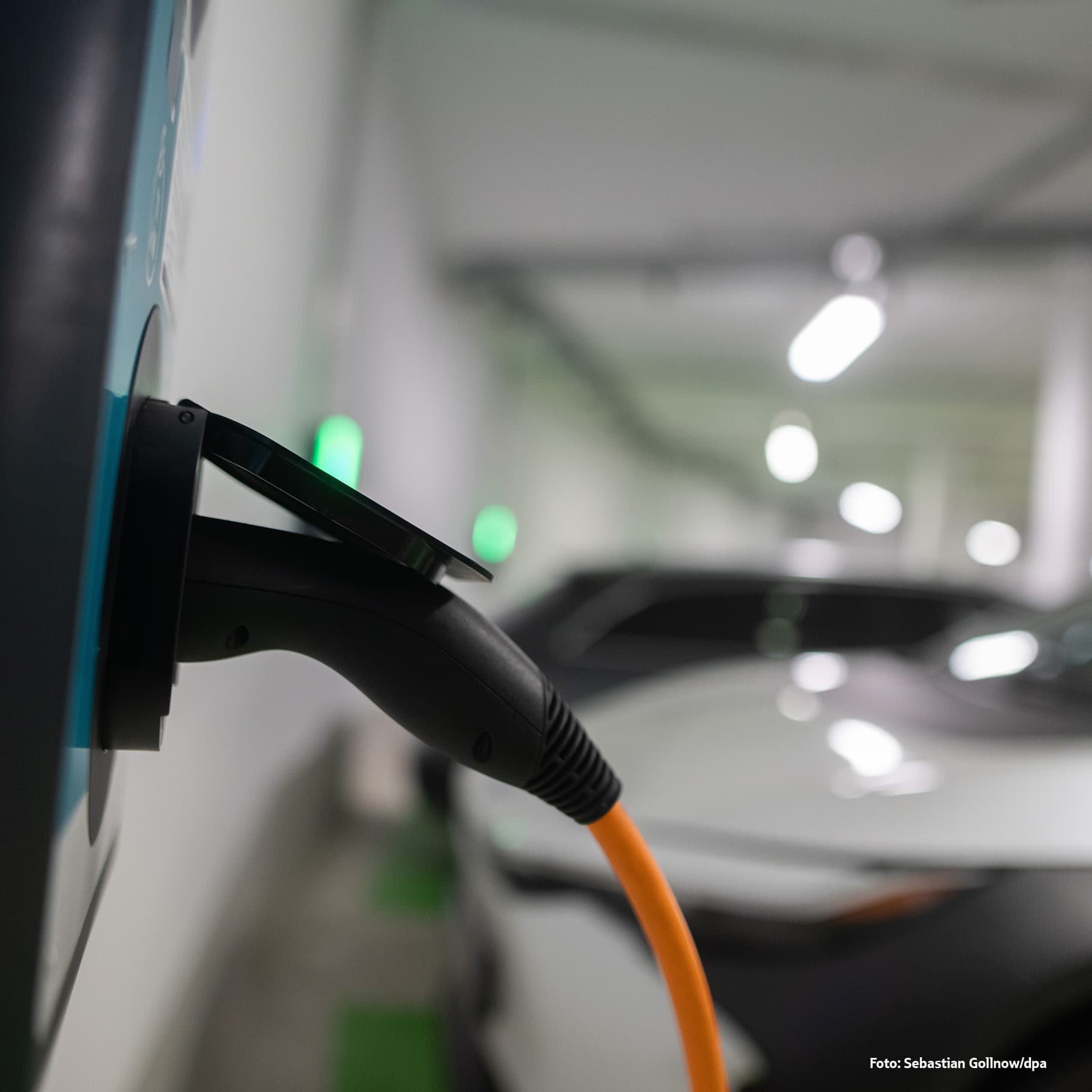 DPA-Bild mit Nahaufnahme eines orangefarbenen Ladekabels, das an eine Ladesäule für Elektroautos angeschlossen ist, mit einem unscharfen Auto und Lichtern im Hintergrund.