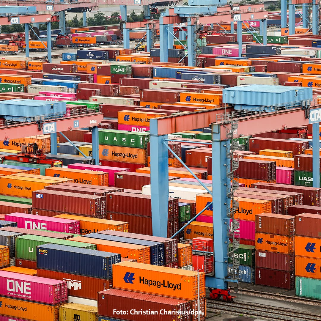 Viele bunte Seecontainer gestapelt in einem Containerterminal mit Krananlagen im Hintergrund.