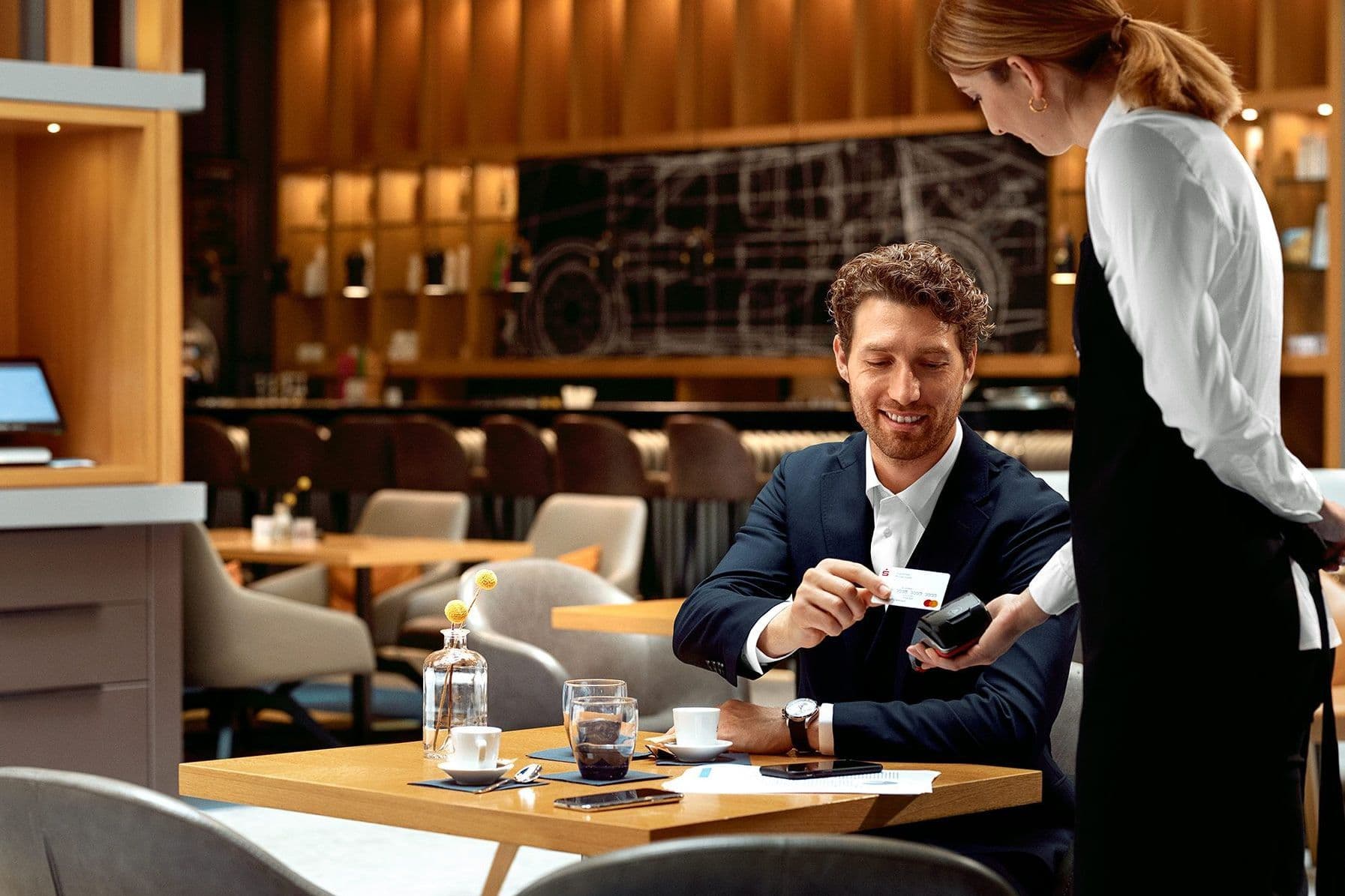 Ein Geschäftsmann mittleren Alters sitzt in einem modernen Restaurant. Er legt lächelnd seine Sparkassenkarte auf ein Kartenlesegerät, welches eine weibliche Servicekraft ihm anreicht.