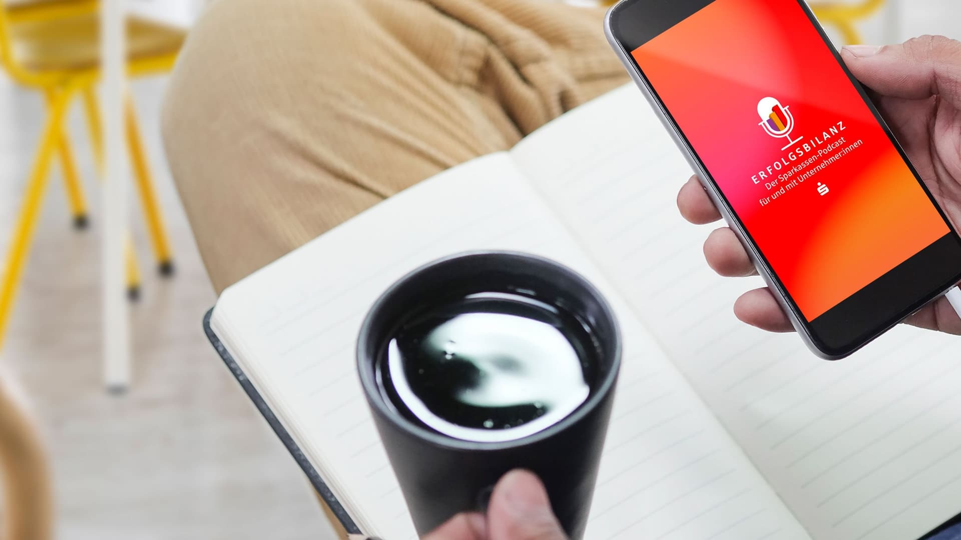 in Notizbuch liegt auf einem Bein.Kaffeetasse in der linken Hand. Smartphone in der rechten Hand. Das Display ist orange mit Logo Erfolgsbilanz.