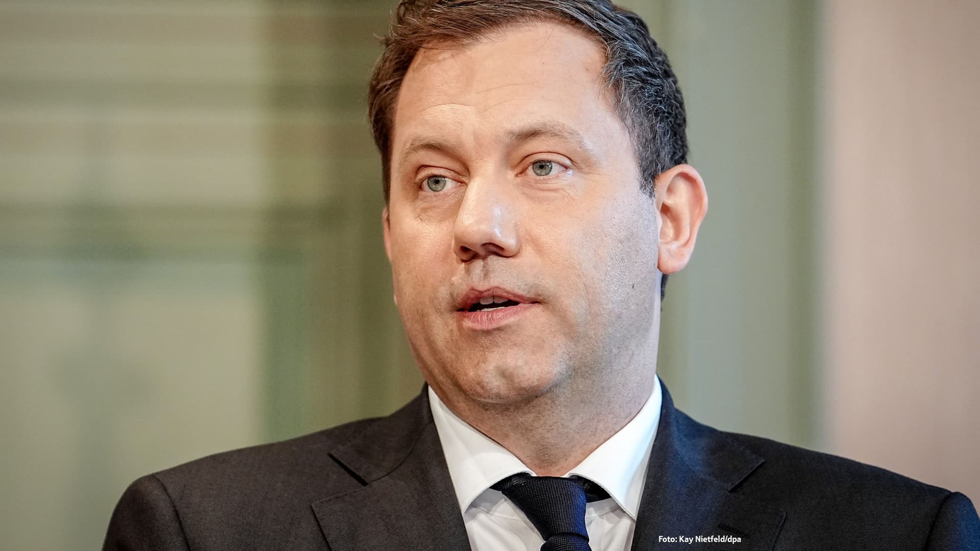 Porträt von Lars Klingbeil im dunklen Anzug mit weißem Hemd und dunkler Krawatte, stehend vor unscharfem, hellgrünlichem Hintergrund.