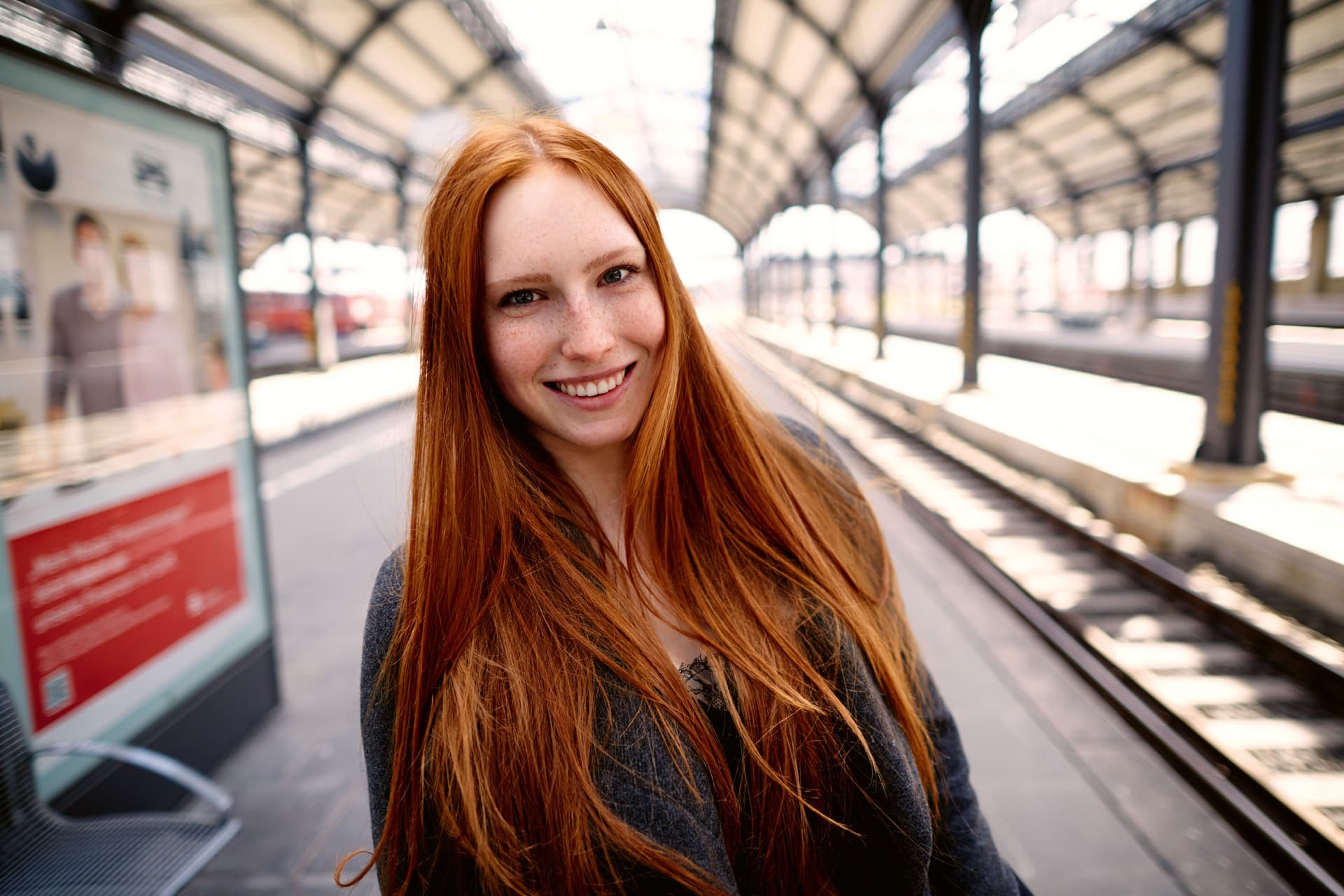 Eine Frau mit langen roten Haaren steht auf einem Bahnsteig.
