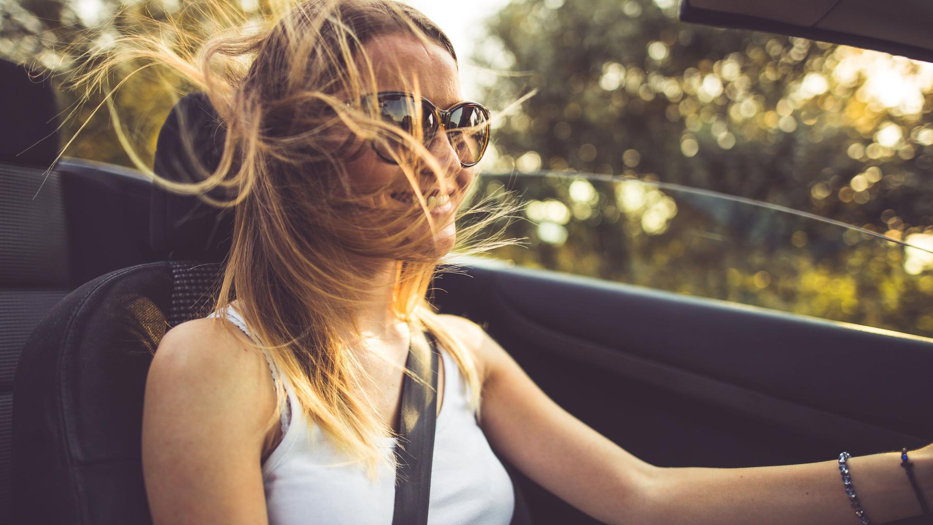 Eine junge, blonde Frau fährt mit wehendem Haar und Sonnenbrille Cabrio.