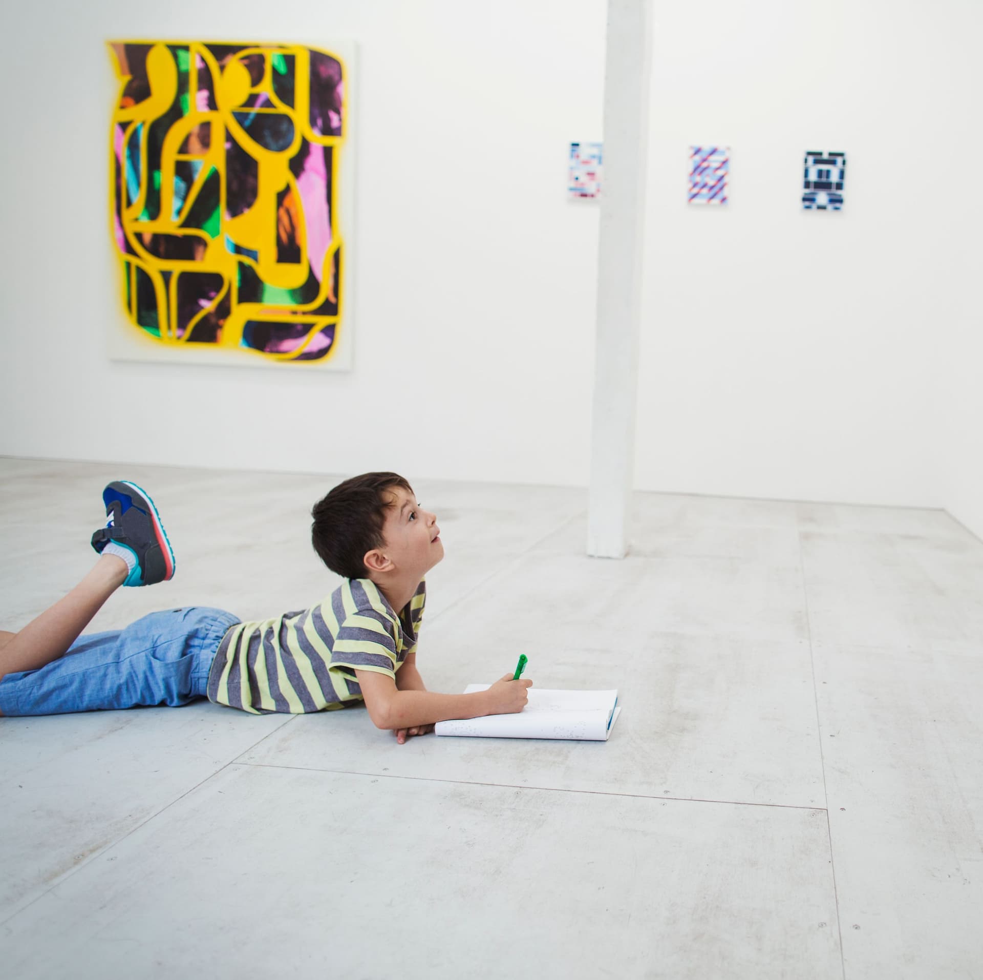 Boy with short black hair lying on floor in art gallery with pen and paper, looking at modern painting.