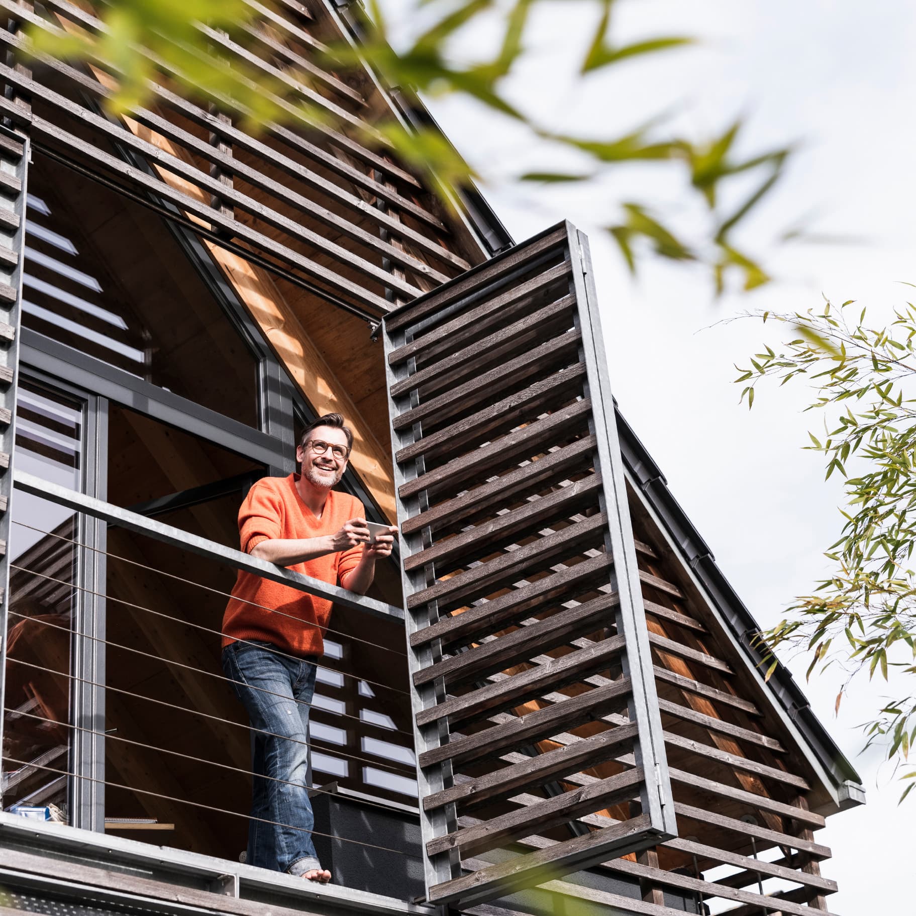 Mann in orangem Pullover steht auf dem Balkon eines modernen Holzhauses; Holzverkleidung dient zur Absicherung des Hauses, rechts Bambuszweige im Vordergrund.