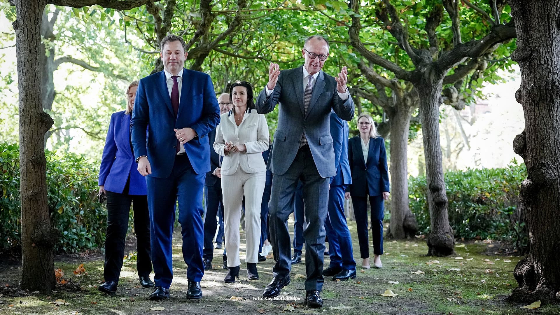 Eine Gruppe von Politikerinnen und Politikern geht im Grünen unter einem Blätterdach entlang. Im Vordergrund spricht Friedrich Merz im grauen Anzug, während Lars Klingbeil im blauen Anzug, Dorothee Bär, Bärbel Bas und weitere Personen folgen.