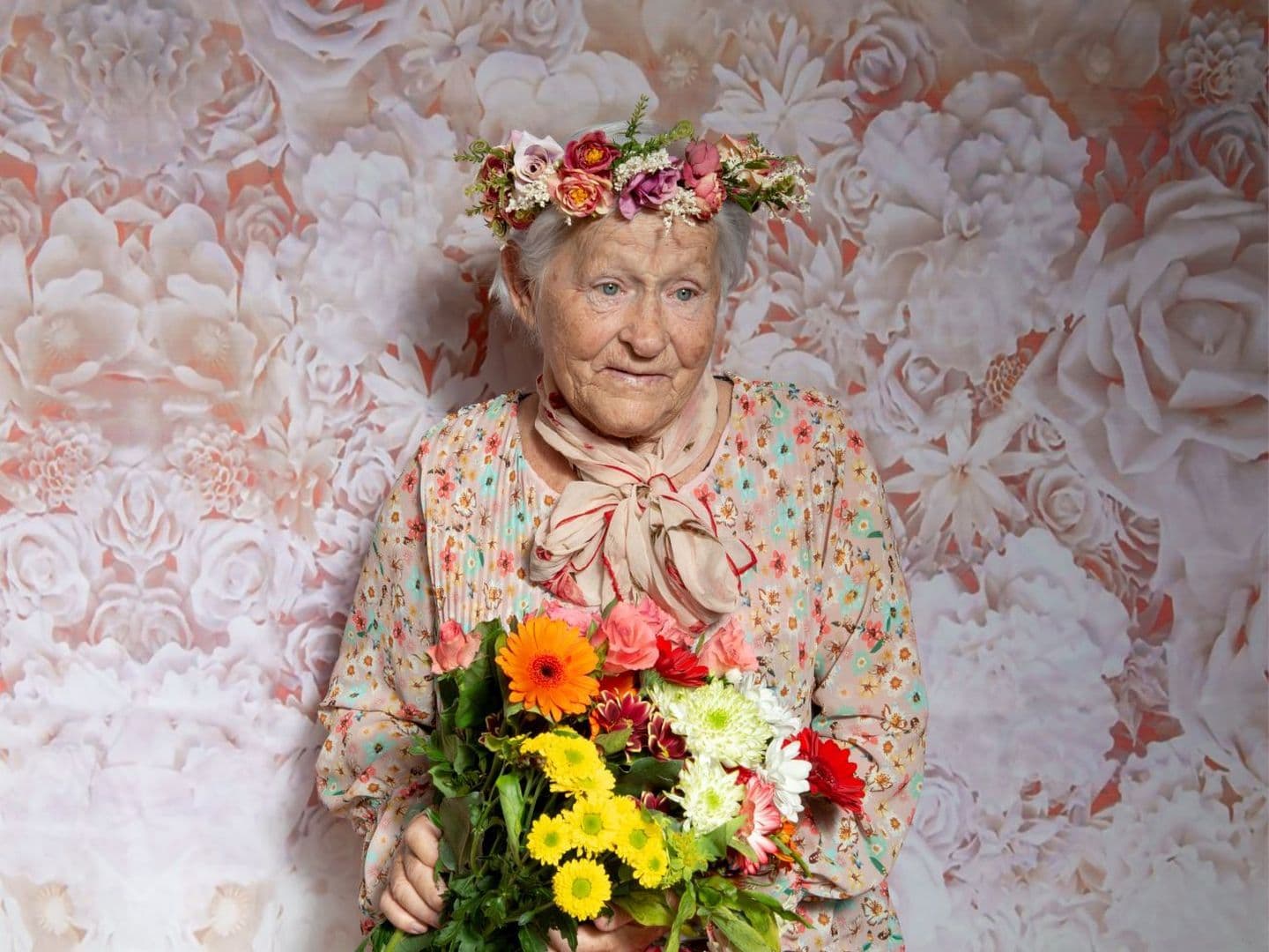 Eine ältere Frau mit weißem Haar steht vor einem pastellfarbenen Hintergrund mit großflächigem Blumenmuster. Sie trägt ein geblümtes Kleid mit Schleifenkragen in Rosa-Beige und eine Krone aus frischen Blumen auf dem Kopf.
