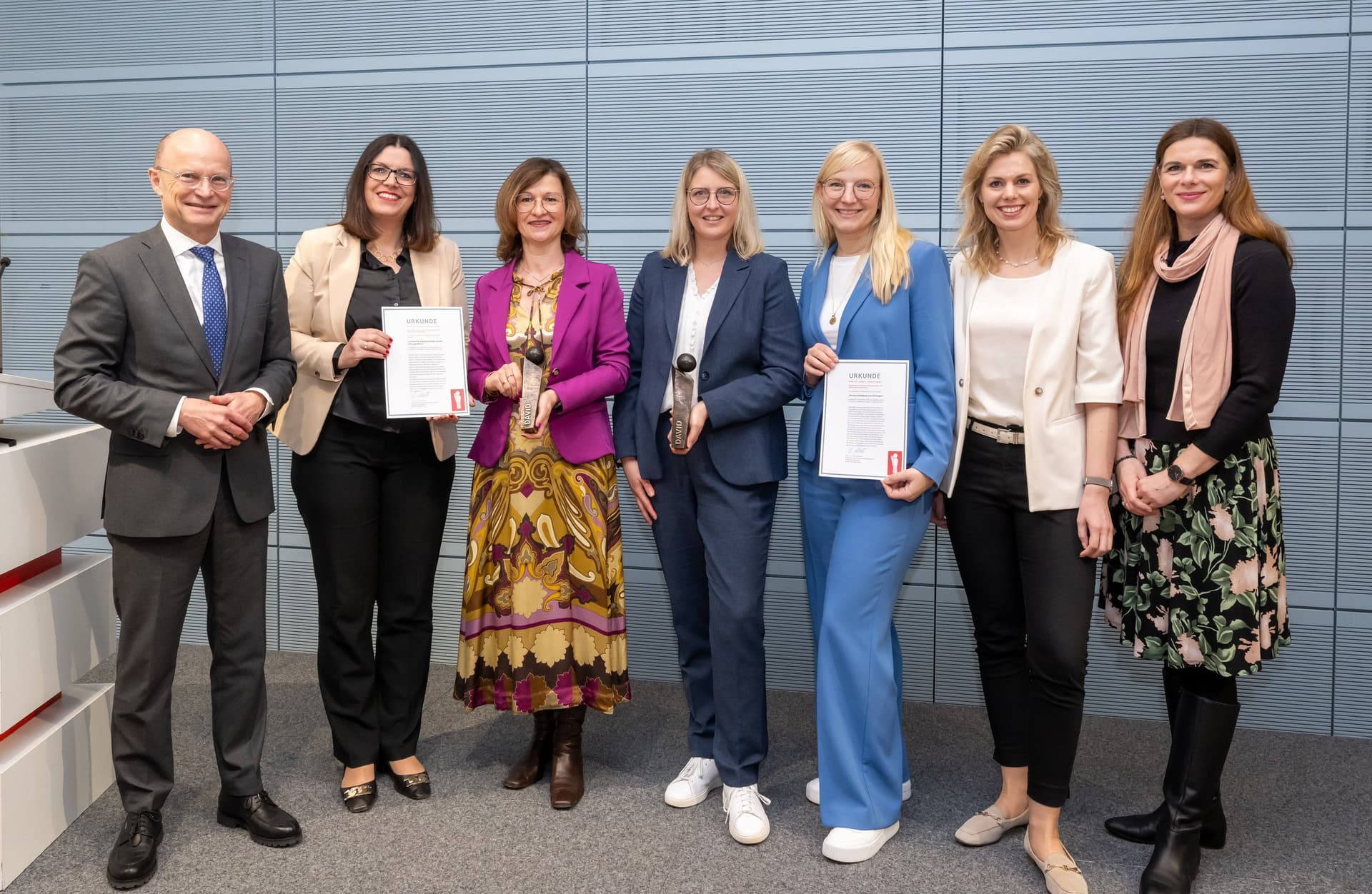 Sieben Personen stehen nebeneinander bei einer Preisverleihung, vier Frauen halten Urkunden und Trophäen, alle lächeln in die Kamera.