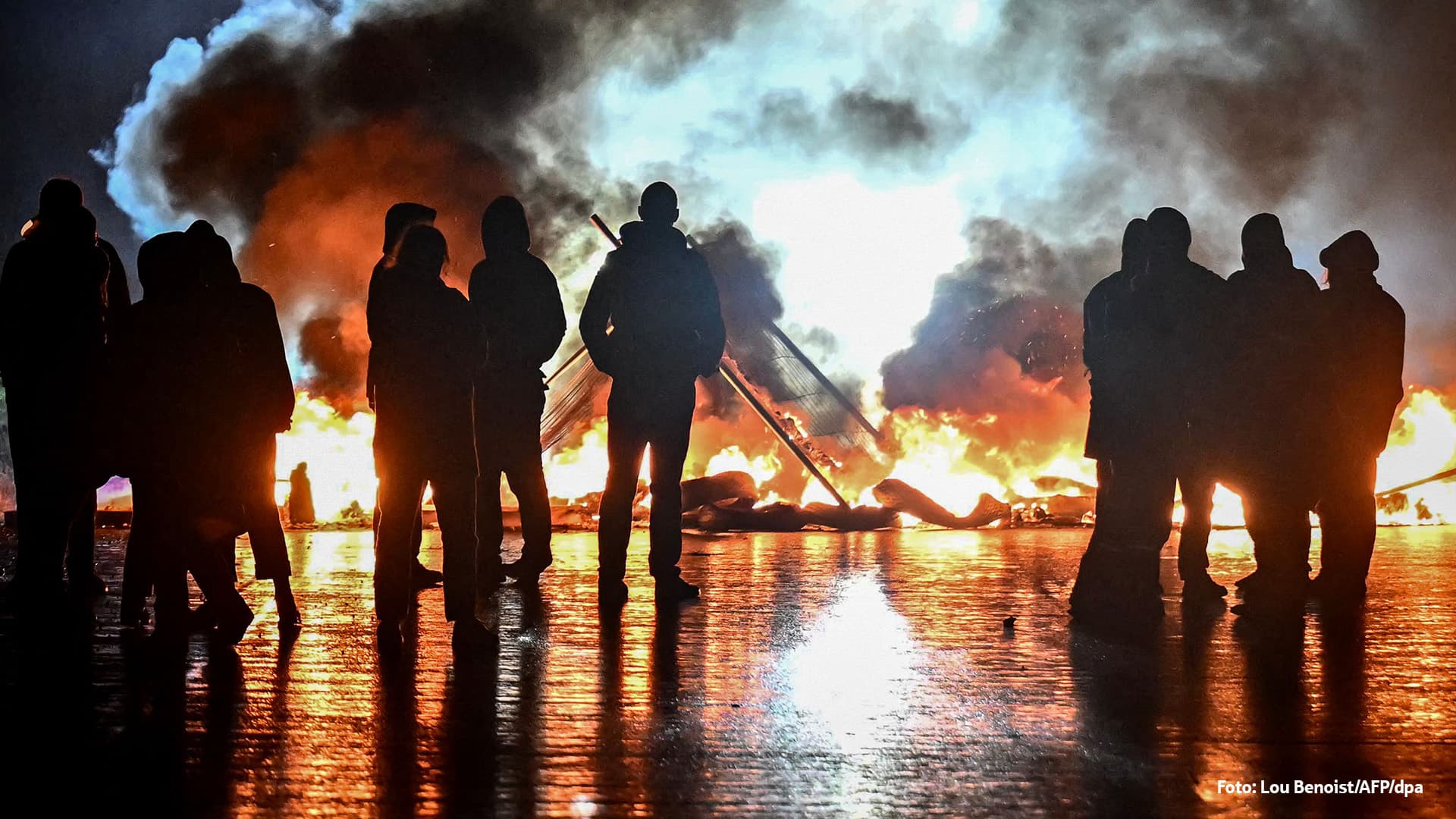 Das Bild zeigt mehrere Menschen, die im Dunkeln vor einem großen Feuer stehen. Die Flammen und dichten Rauchwolken erhellen die Szene, während die Personen nur als Schattenrisse erkennbar sind.