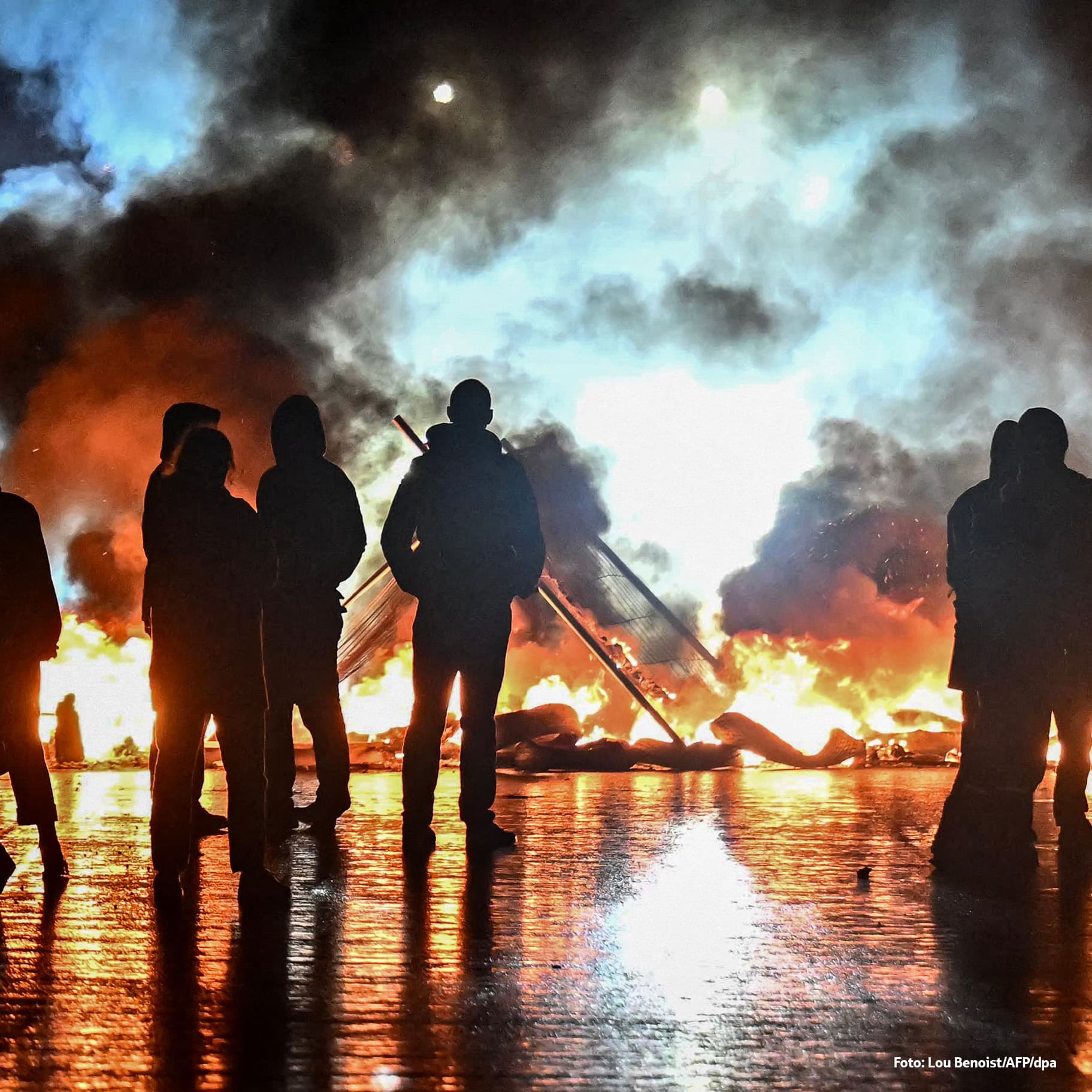 Das Bild zeigt mehrere Menschen, die im Dunkeln vor einem großen Feuer stehen. Die Flammen und dichten Rauchwolken erhellen die Szene, während die Personen nur als Schattenrisse erkennbar sind.