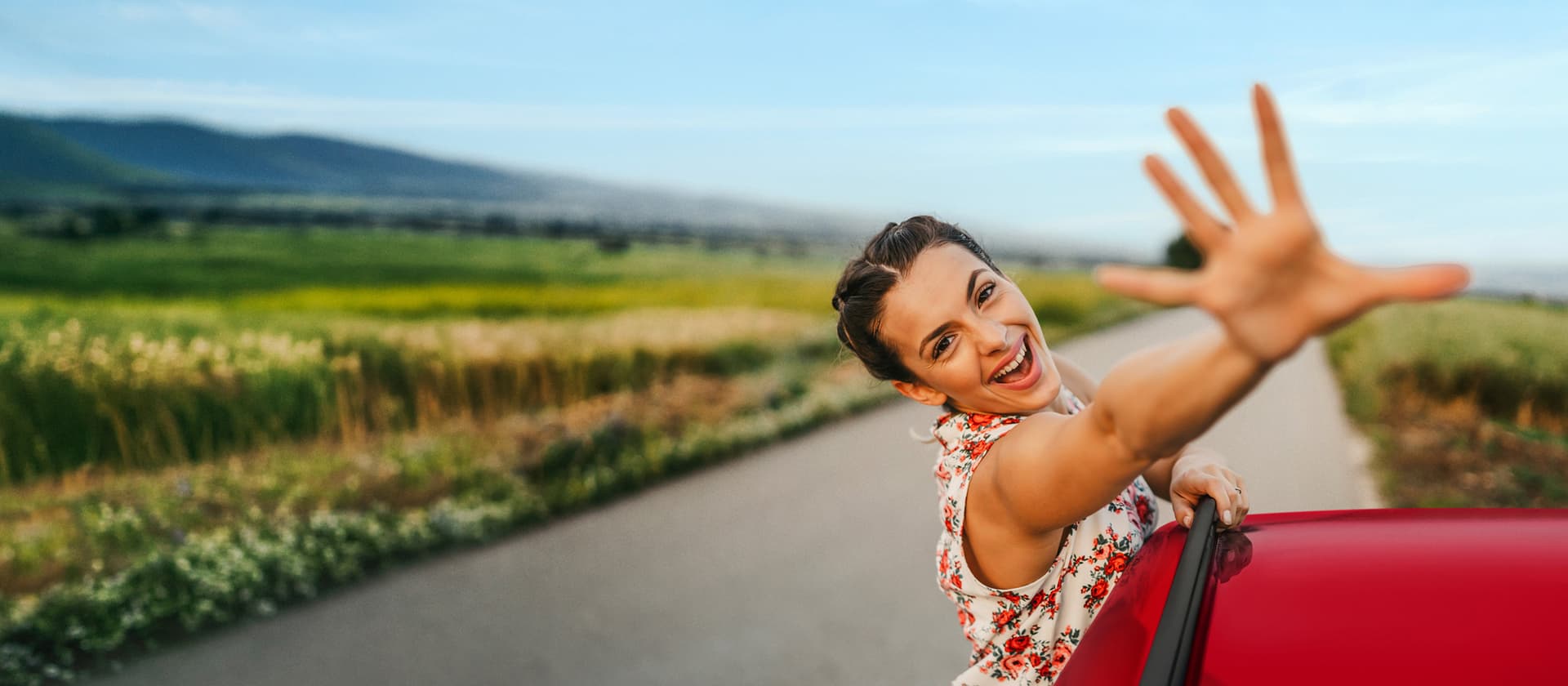Eine lächelnde Frau lehnt sich aus einem roten Auto, streckt ihre Hand aus und genießt die Fahrt auf einer Landstraße mit grünen Feldern und Bergen im Hintergrund.