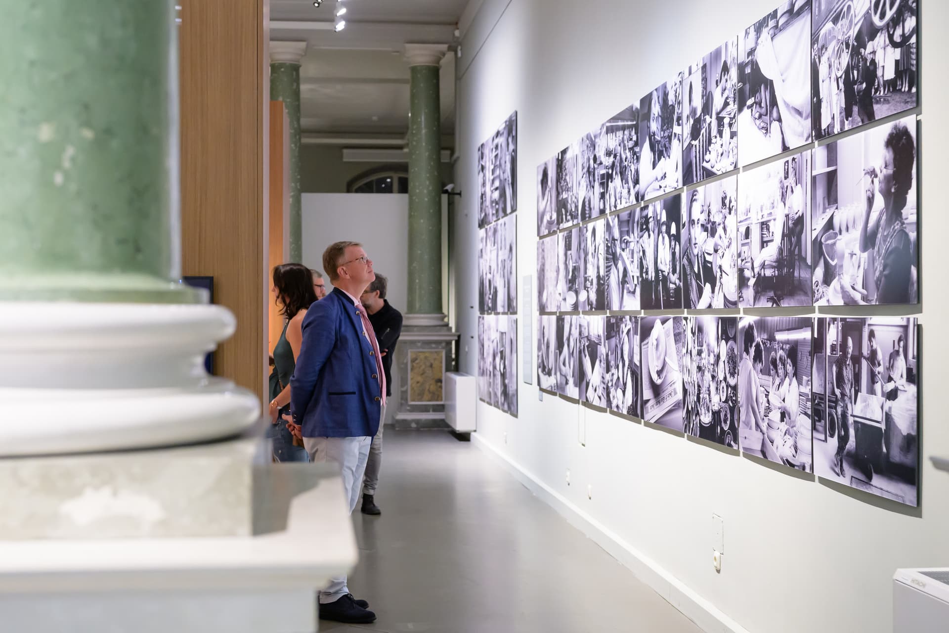 Mehrere Personen betrachten in einer hellen Ausstellungshalle eine Wand mit zahlreichen schwarz-weißen Fotografien, die in einem Raster angeordnet sind. Im Vordergrund steht ein Mann in blauem Sakko, der die Bilder studiert.
