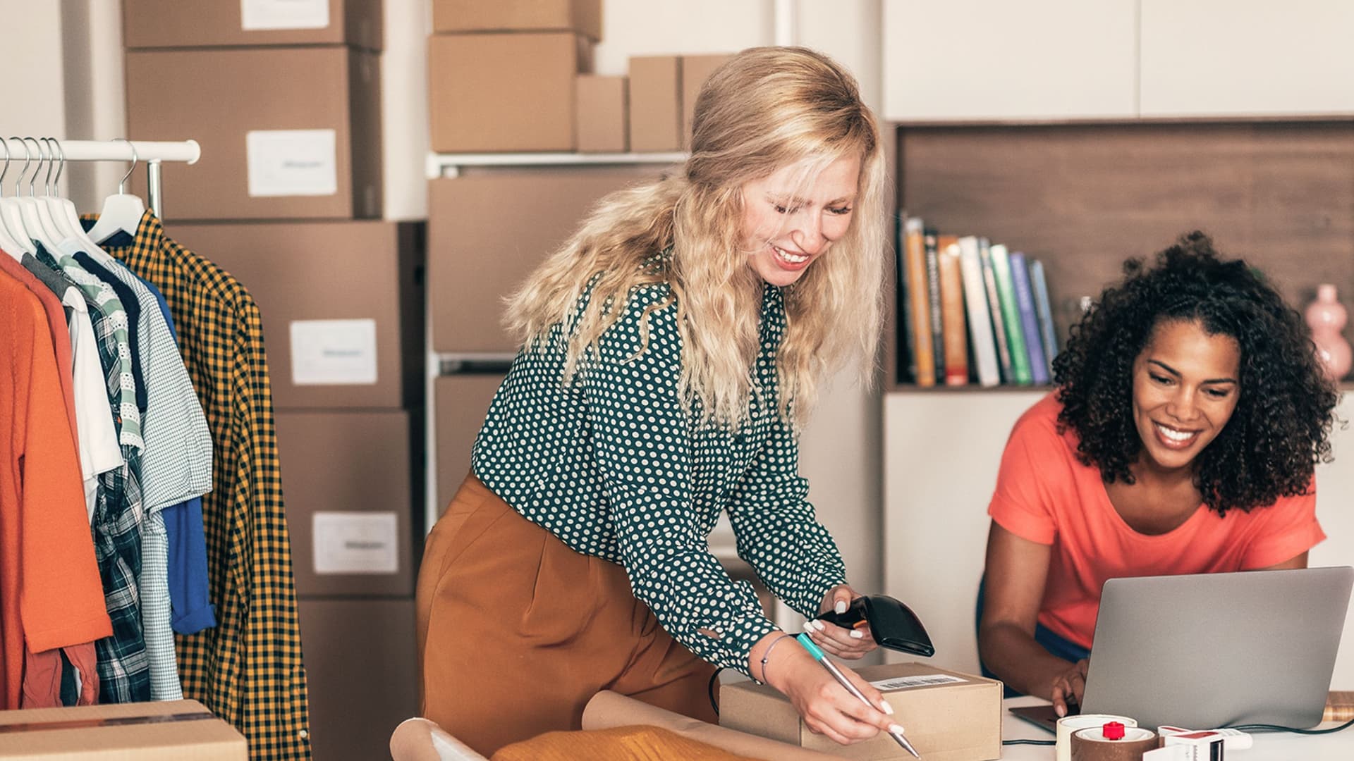 Zwei Frauen arbeiten in einem kleinen Online-Shop: Eine scannt ein Paket, die andere sitzt am Laptop. Im Hintergrund sind Kartons, Kleidung und Bücher zu sehen.