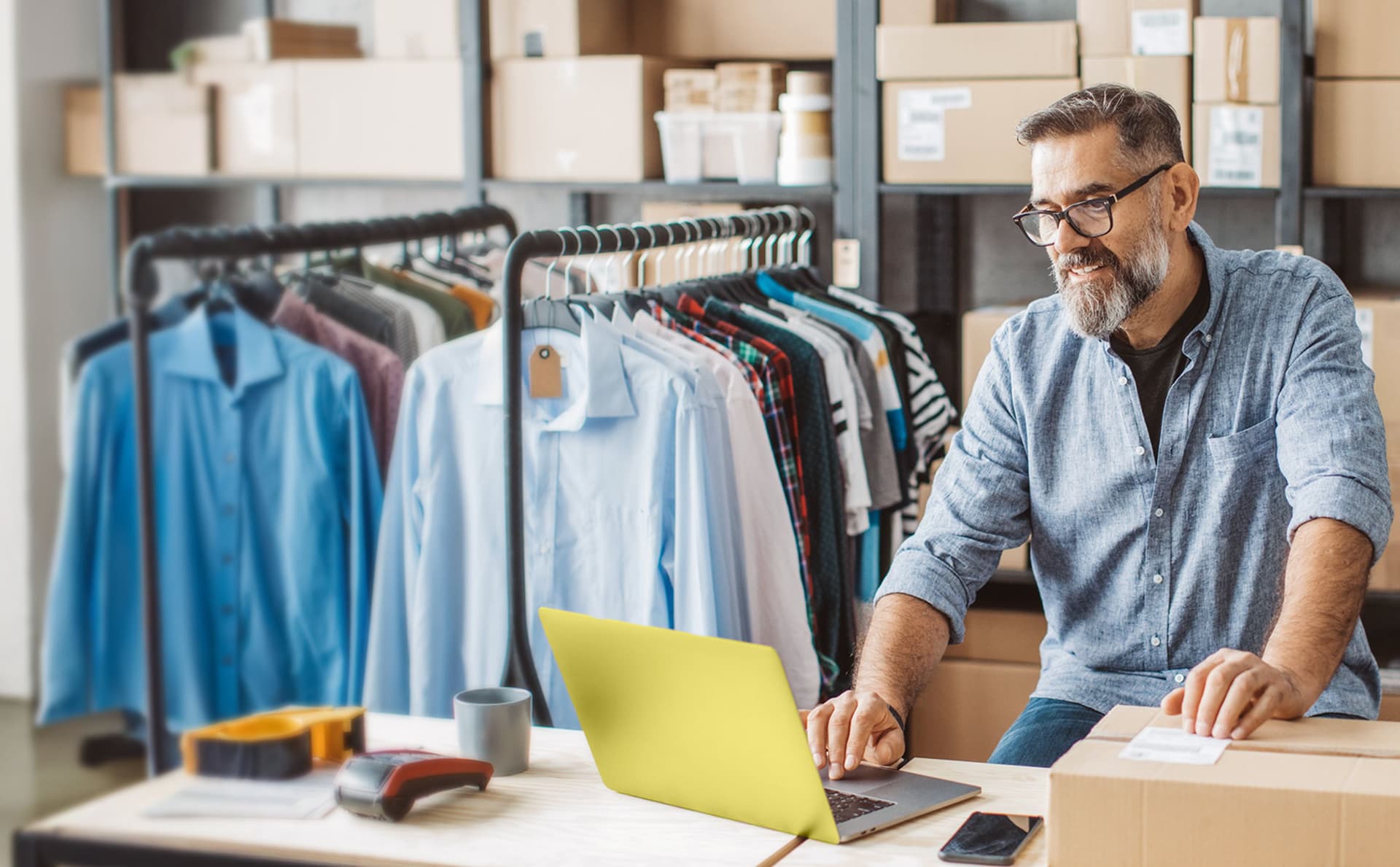 Ein Mann mit Brille und Bart arbeitet an einem Laptop in einem kleinen Bekleidungslager. Hinter ihm hängen Hemden und T-Shirts auf einer Kleiderstange, während um ihn herum Kartons und Verpackungsmaterialien zu sehen sind.