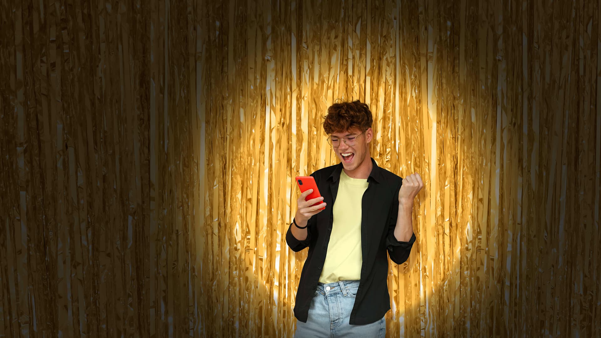 Junger Mann mit Smartphone steht jubelnd vor goldenem Glitzervorhang im warmen Scheinwerferlicht.