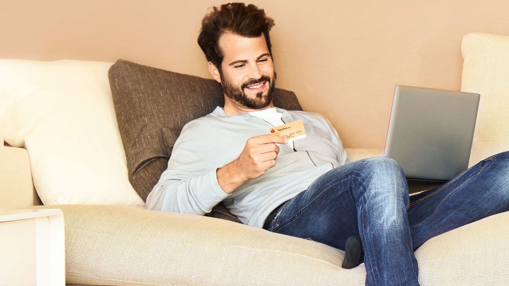 Mann sitzt mit einer Kreditkarte und seinem Laptop auf dem Schoß auf der Couch.