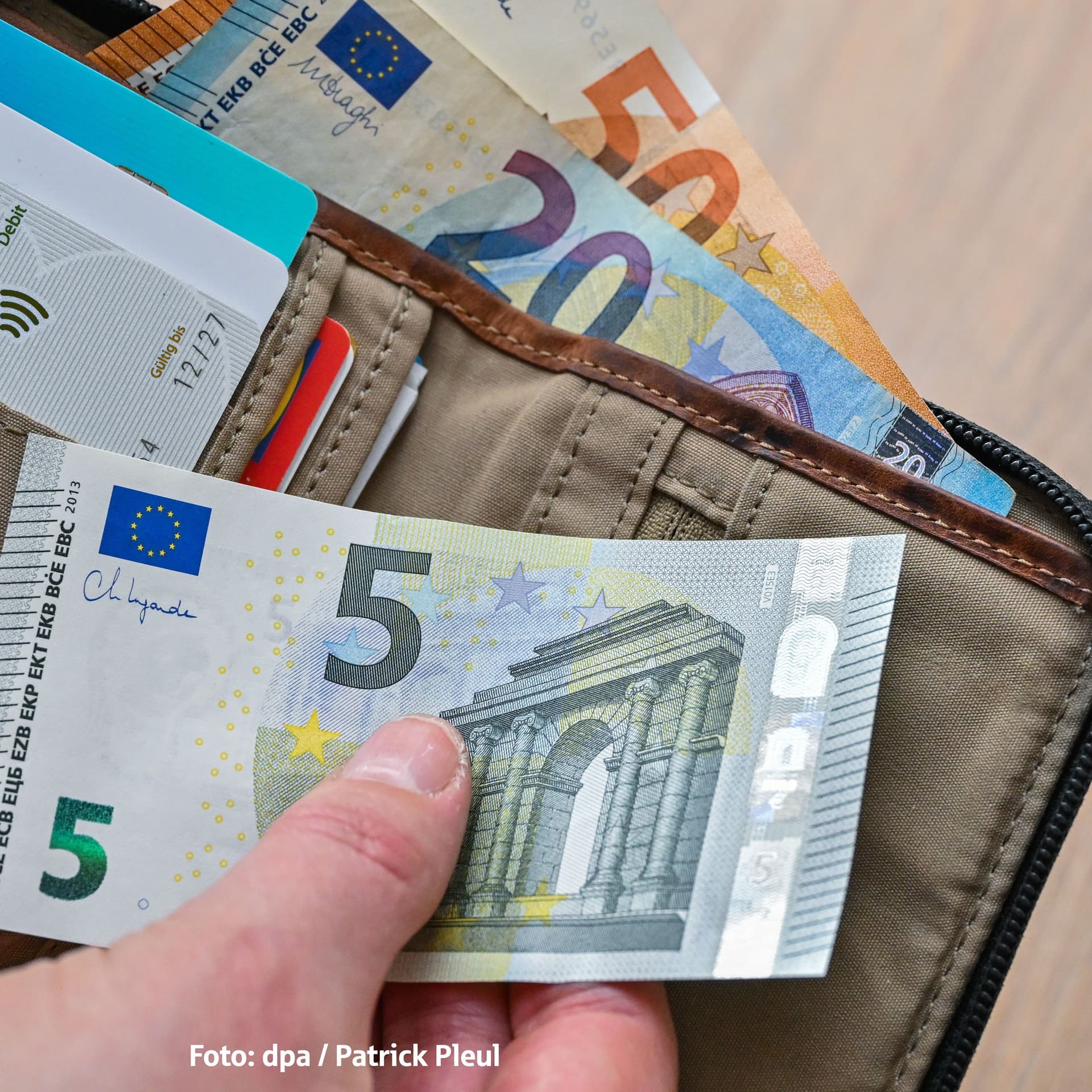 Eine Hand hält einen 5-Euro-Schein. Dahinter liegt ein Geldbeutel mit einem 20-Euro Schein und einem 50-Euro Schein.