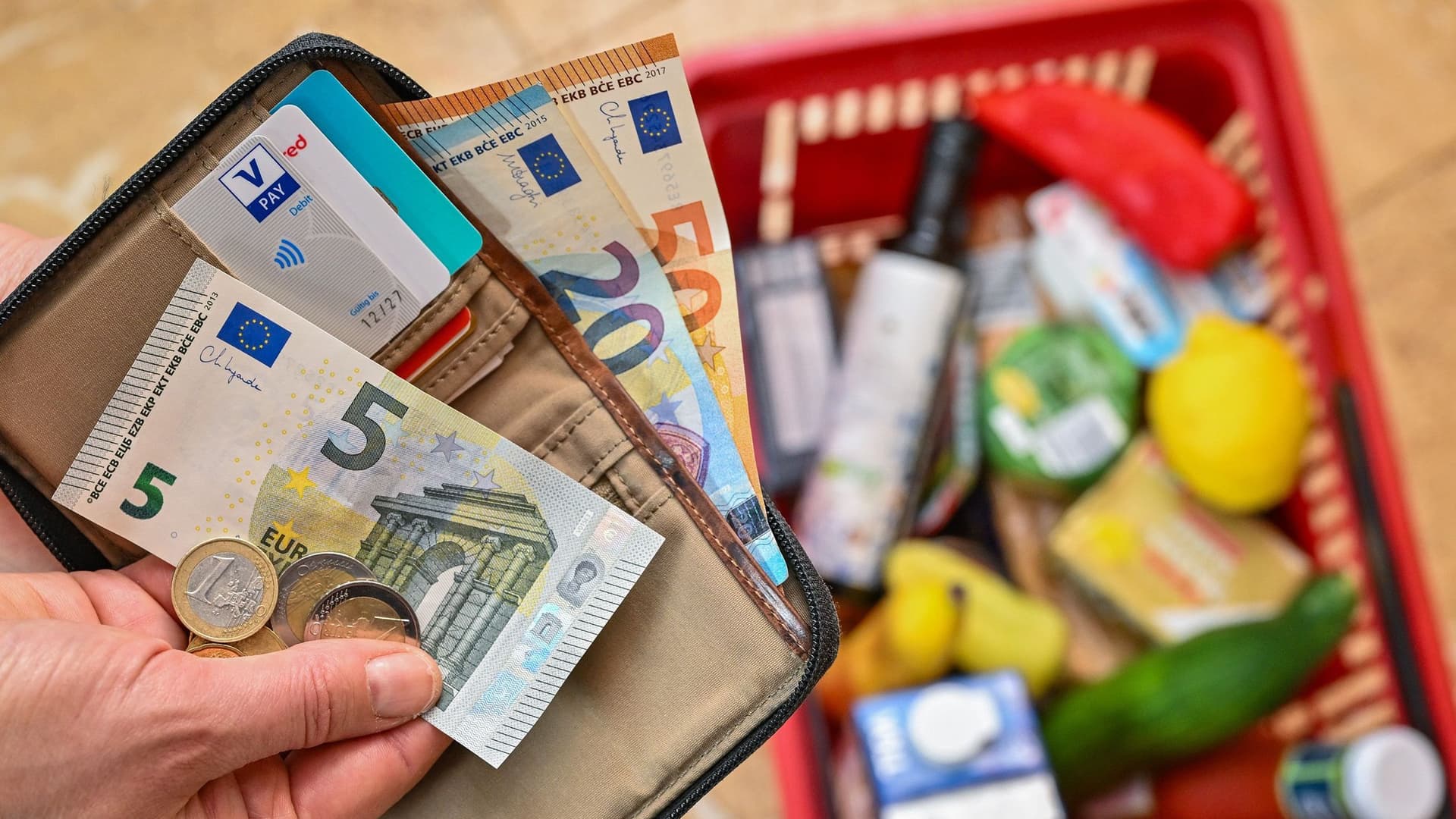 Eine Hand hält einen Geldbeutel mit Geld. Im Hintergrund ist ein Einkaufskorb zu sehen.