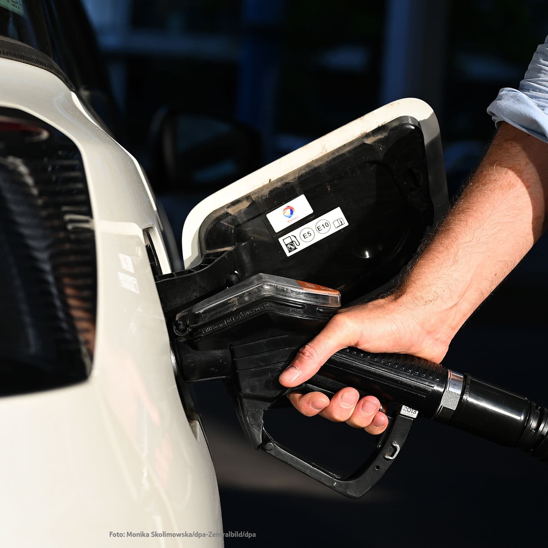 Ein Arm führt eine Zapfpistole in den geöffneten Tankstutzen eines weißen Autos, während das Heck mit Rücklicht im Bildrand sichtbar ist.