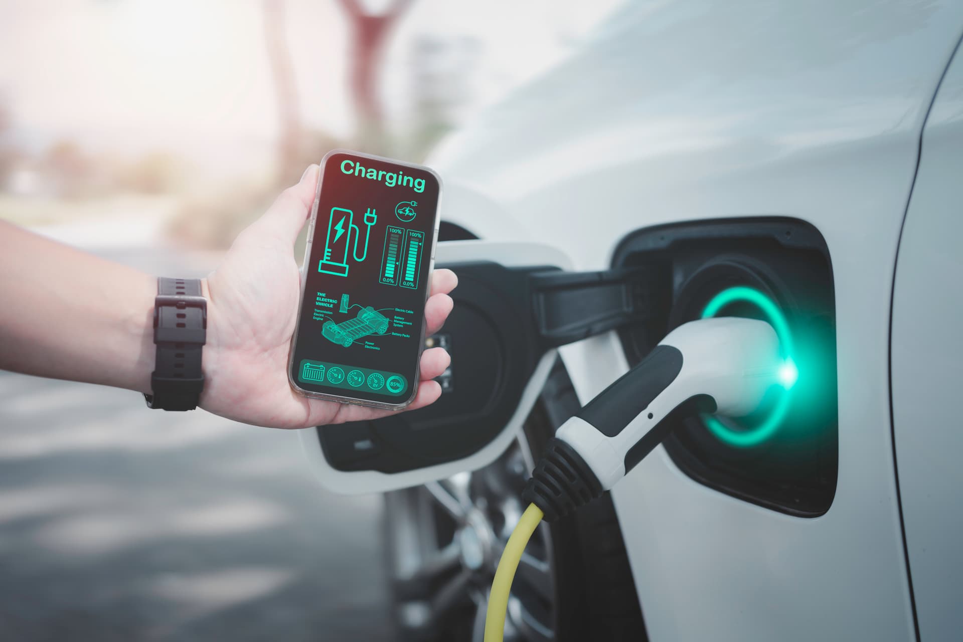Eine Hand hält ein Smartphone mit Ladeanzeige neben einem Elektroauto, das gerade an einer Ladestation angeschlossen ist.