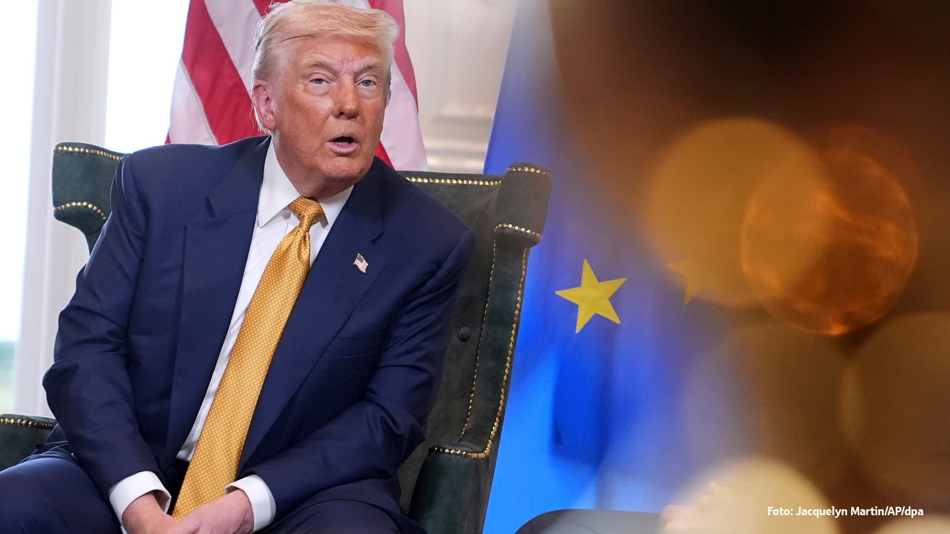 Donald Trump sitzt auf einem Stuhl vor den Flaggen der USA und der Europäischen. Die Aufnahme ist teilweise von verschwommenen Lichtreflexen im Vordergrund überlagert.