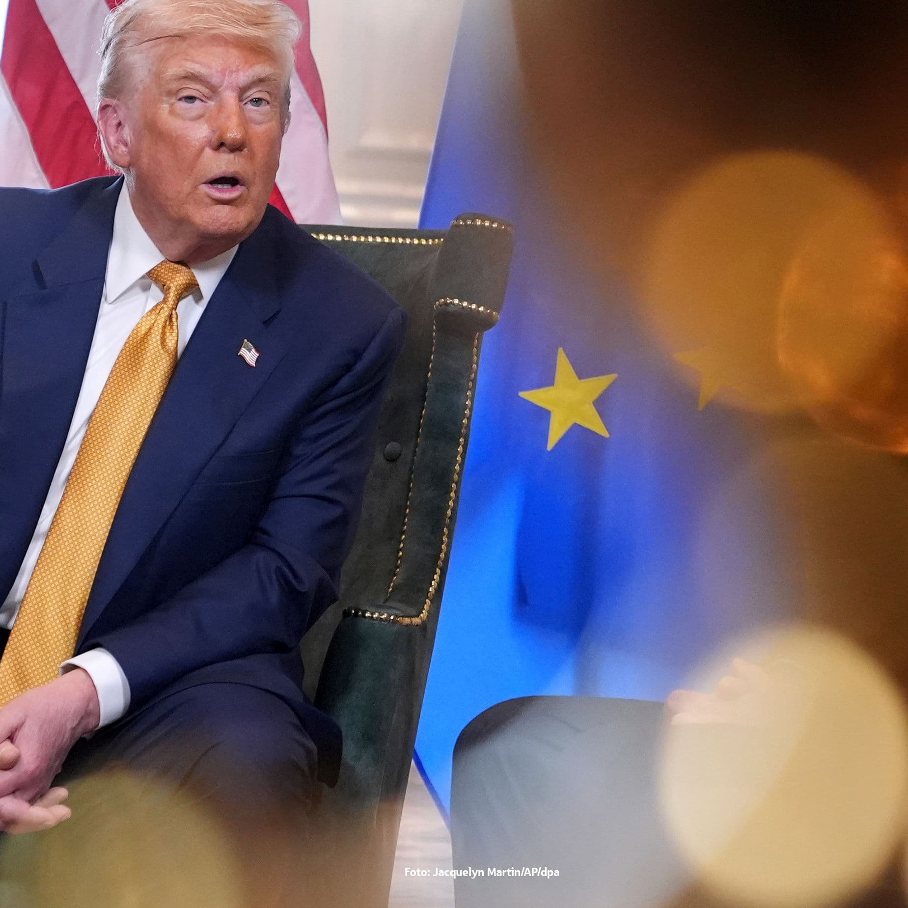 Donald Trump sitzt auf einem Stuhl vor den Flaggen der USA und der Europäischen. Die Aufnahme ist teilweise von verschwommenen Lichtreflexen im Vordergrund überlagert.