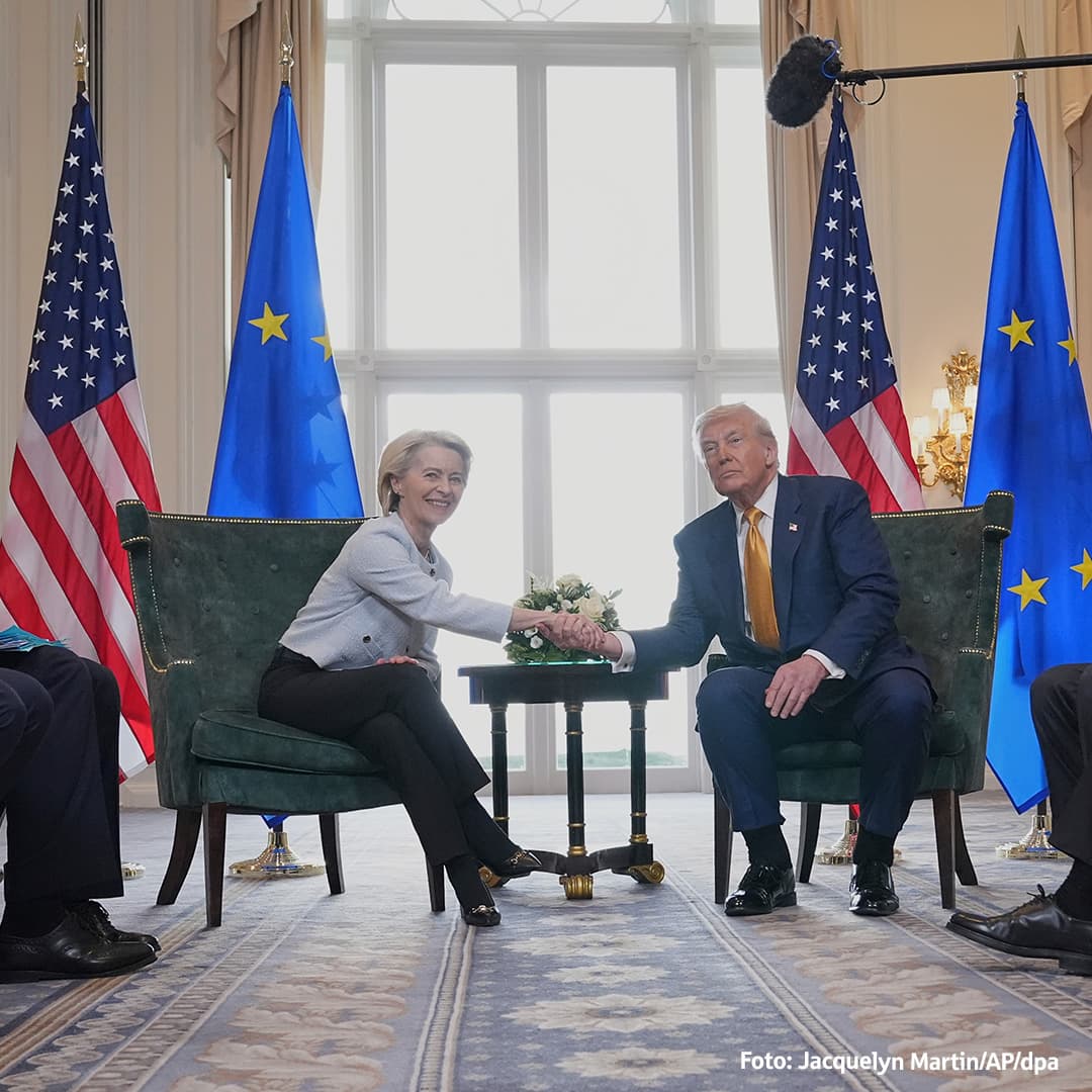  Ursula von der Leyen und Donald Trump geben sich bei einem Treffen die Hand, im Hintergrund stehen EU- und US-Flaggen.