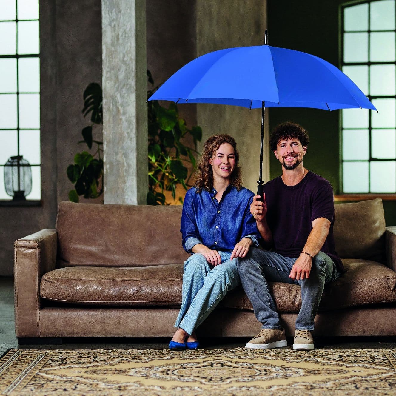 Ein Paar sitzt lächelnd auf einem braunen Ledersofa in einem wohnlich gestalteten Raum und hält gemeinsam einen großen, blauen Regenschirm über sich. Der Boden im Hintergrund ist mit Wasser überflutet