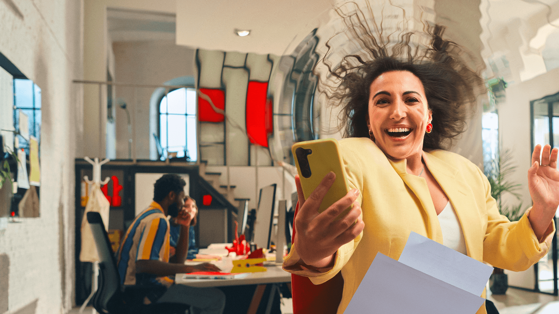 Eine fröhliche Frau macht ein Selfie im Büro, während ihre Haare und Papiere durch die Luft wirbeln.