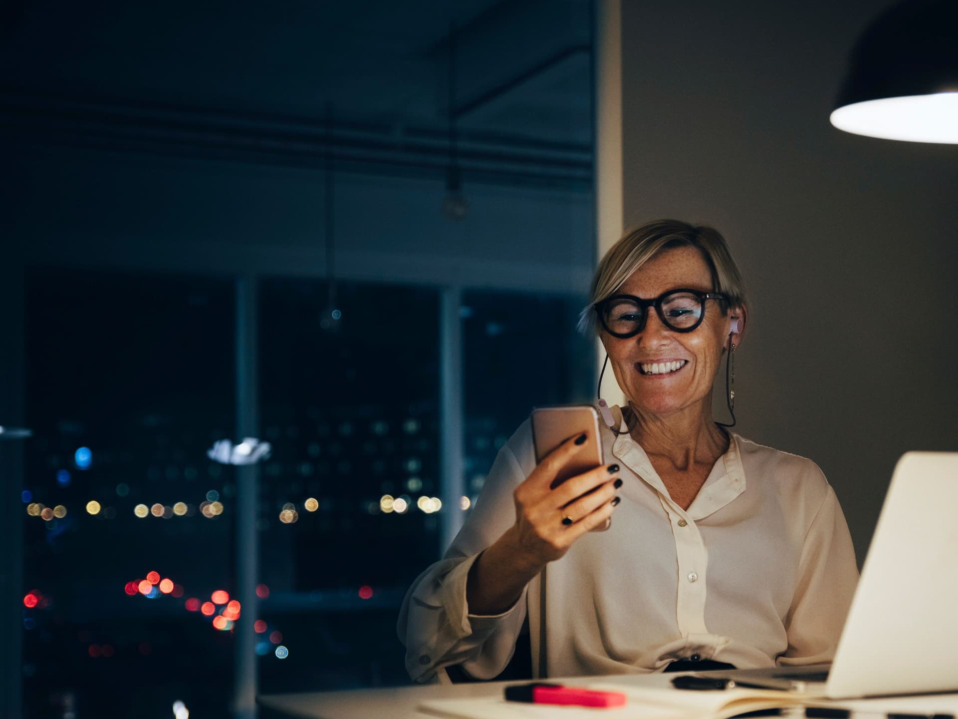 Lächelnde Frau sitzt mit Smartphone in der Hand an einem Schreibtisch. Ihr Laptop steht auf dem Tisch. Durch das Fenster sieht man Lichter einer Stadt, es ist dunkel.
