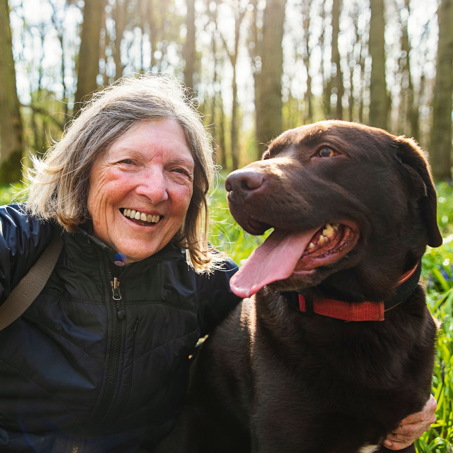 Eine Frau Dame macht ein Selfie mit einem Hund auf einer Wiese. Im Hintergrund scheint die Sonne durch die Bäume.