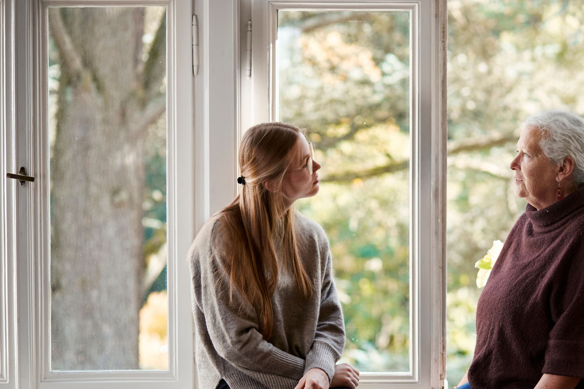 Ältere Frau und junge Frau sitzen sich zugewandt auf der Fensterbank. Hinter ihnen sind Altbaufenster mit Blick ins Grüne.