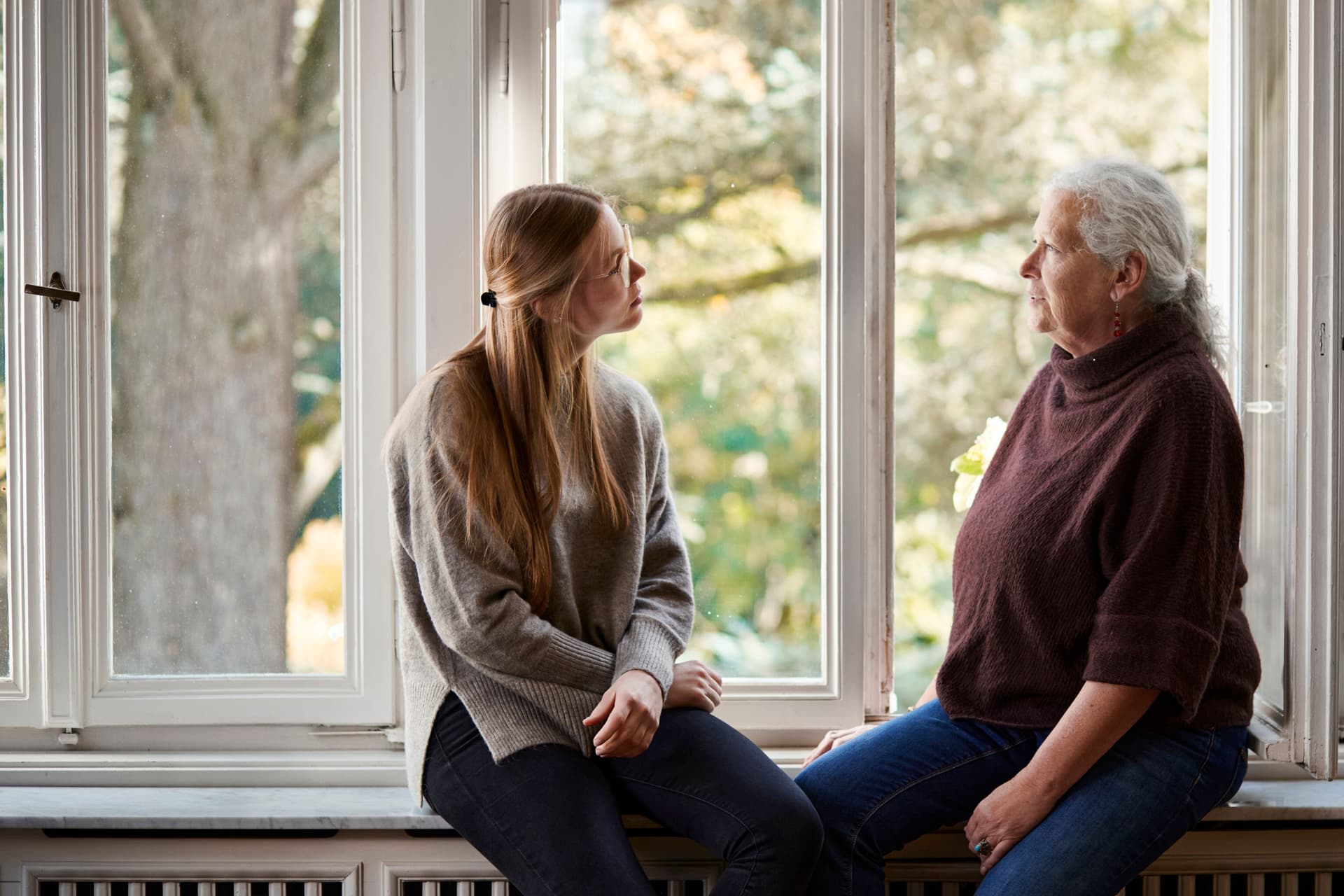 Ältere Frau und junge Frau sitzen sich zugewandt auf der Fensterbank. Hinter ihnen sind Altbaufenster mit Blick ins Grüne.