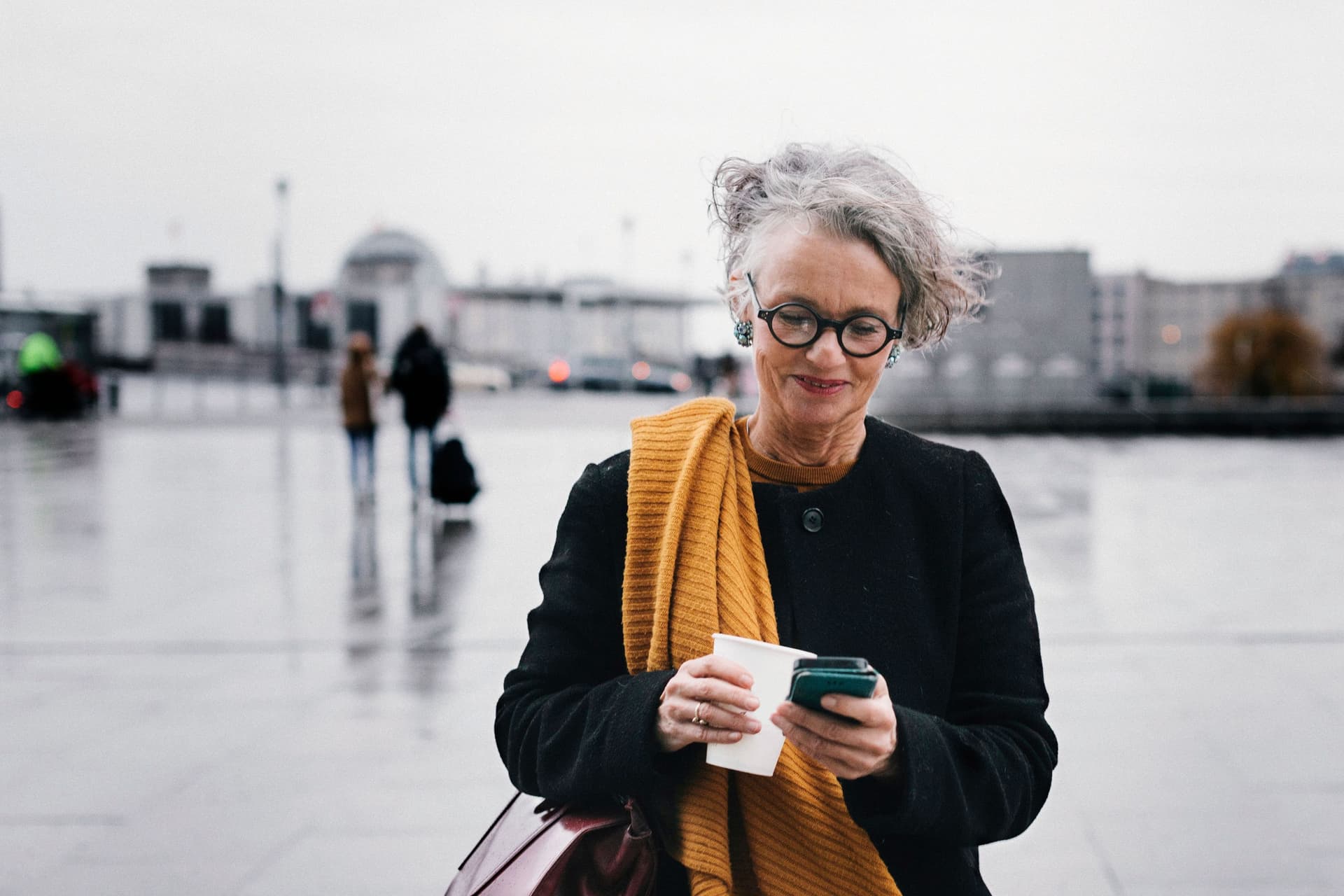 Eine ältere Frau steht mit einem Smartphone und einem Kaffeebecher in der Hand auf einem Platz. Sie schreibt und guckt auf ihr Handy.