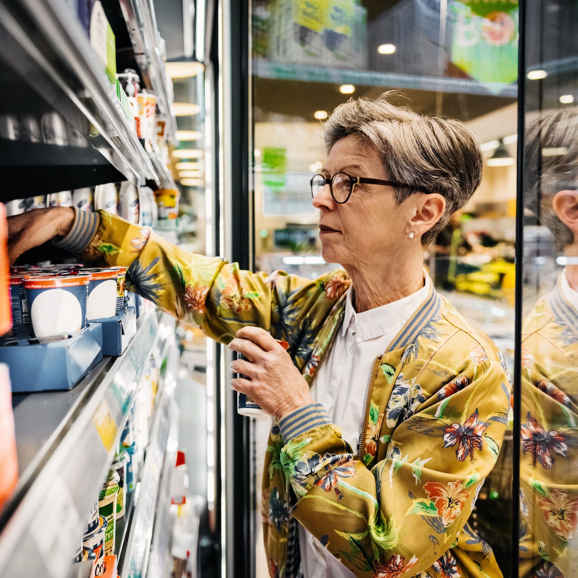 Eine ältere Frau im Supermarkt, die Lebensmittel aus einem Kühlschrank herausholt.